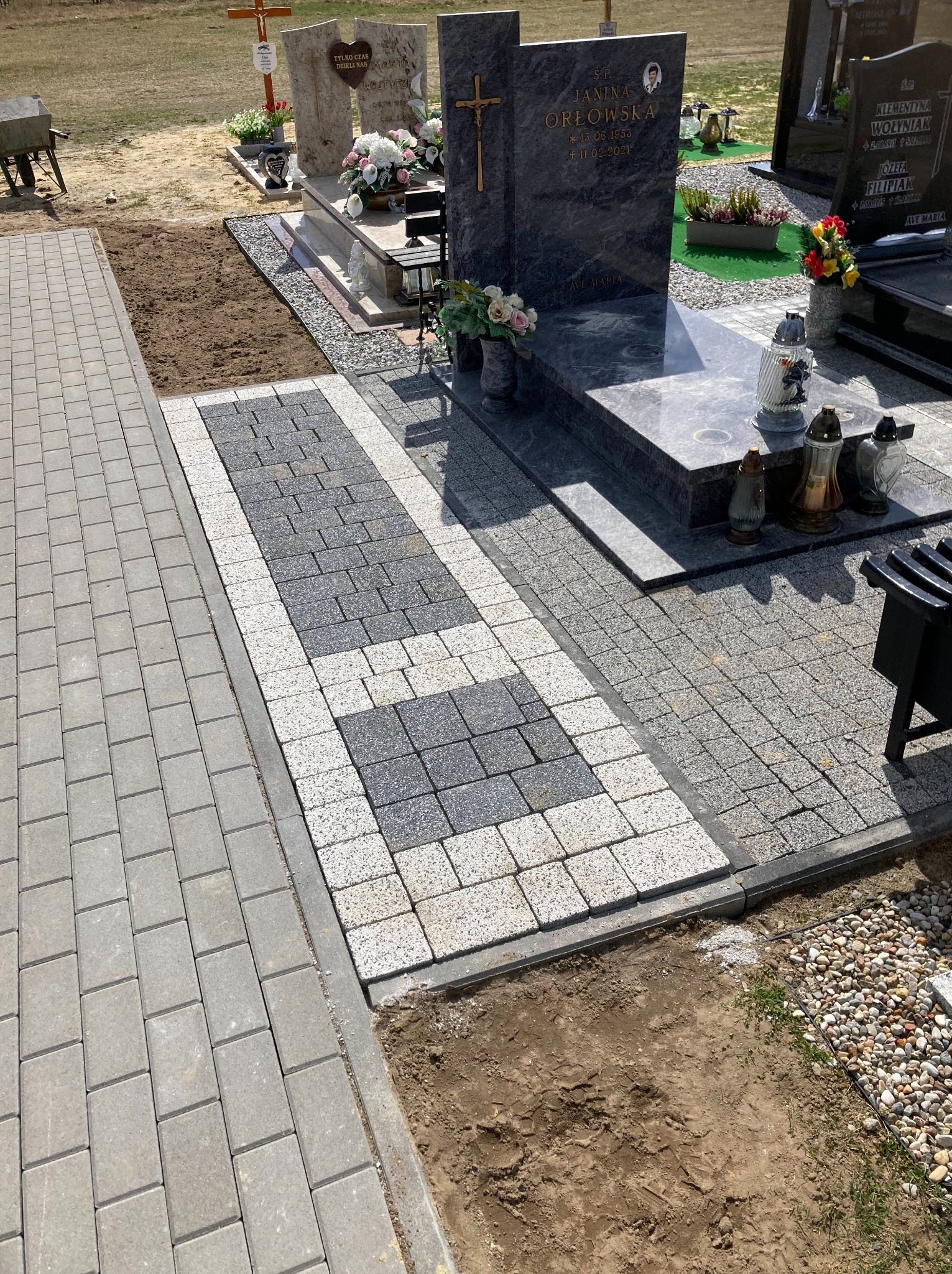 Ścieżka z szarej i białej kostki brukowej na cmentarzu, prowadząca do nagrobków; widoczne świeże prace ziemne wokół.