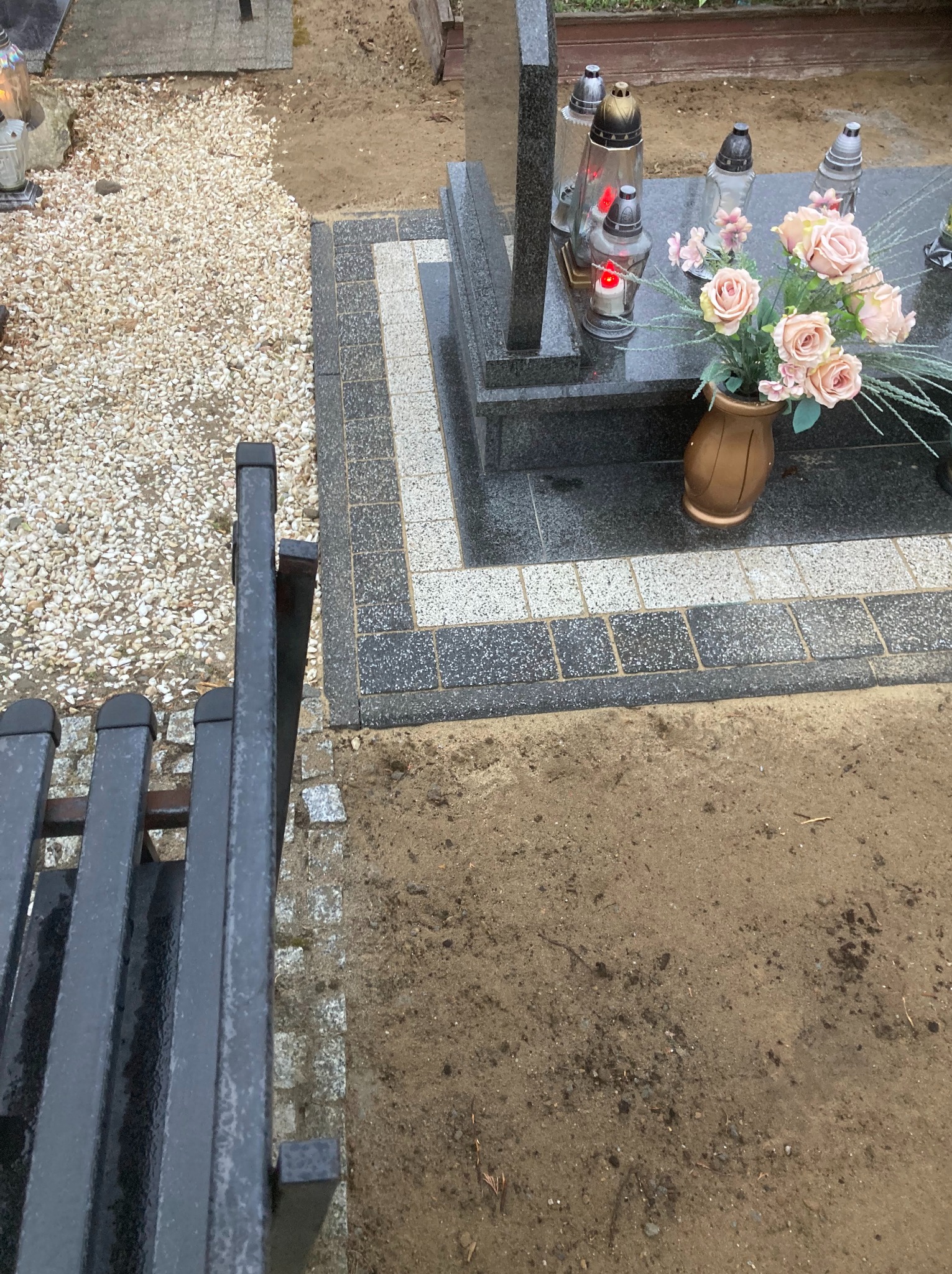 Granitowy nagrobek z zapalonymi zniczami i bukietem róż, otoczony obrzeżem z kostki brukowej w dwóch odcieniach szarości, widok z boku ławki.
