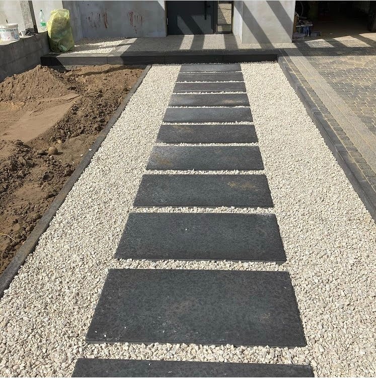 Ścieżka z prostokątnych płyt z ciemnego kamienia ułożona na jasnym żwirze, prowadząca do wejścia budynku, z widocznym fragmentem rabaty ziemnej po lewej stronie.