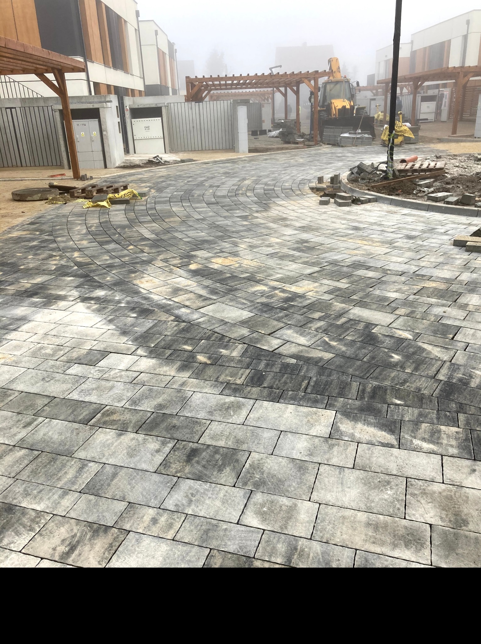 Świeżo ułożona, wilgotna nawierzchnia z szarej kostki brukowej w kształcie łuku, z widocznym w tle placem budowy, pergolami i koparką we mgle.