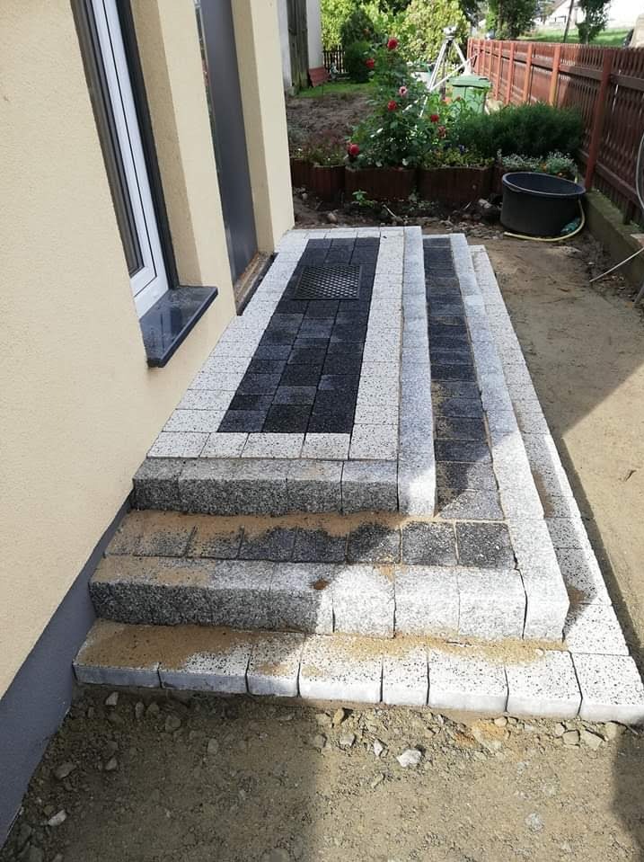 Schody wejściowe do domu wyłożone kostką brukową w odcieniach szarości i czerni z kratką ściekową, widoczne ślady piasku na powierzchni, tło z zielenią ogrodową i elementami drewnianego ogrodzenia.