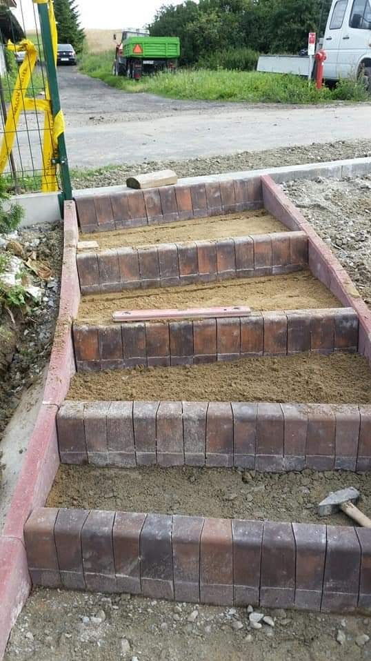 Trzy stopnie schodów z brązowej kostki klinkierowej w trakcie budowy, wypełnione piaskiem, poziomica leży na drugim stopniu, w tle widoczny traktor i samochód osobowy.