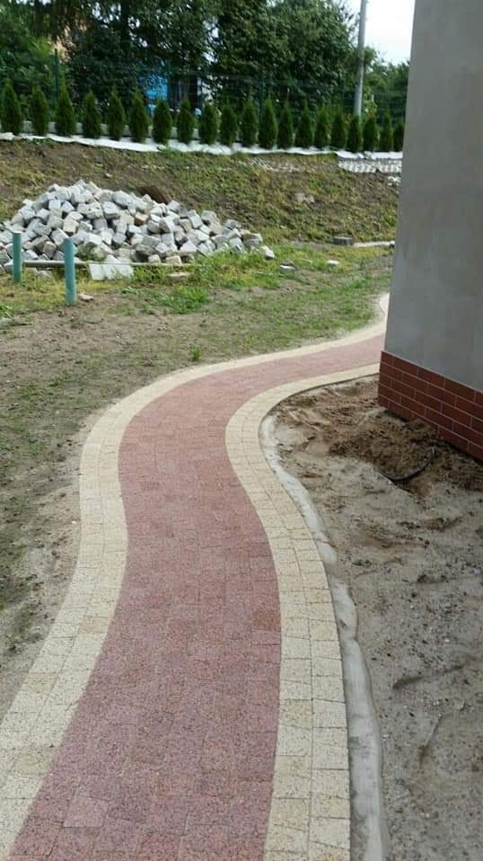 Kręta ścieżka z czerwonej kostki brukowej z jasnymi obrzeżami, prowadząca obok budynku w trakcie budowy, z stertą kostki w tle i rzędem małych drzewek na wzgórzu.