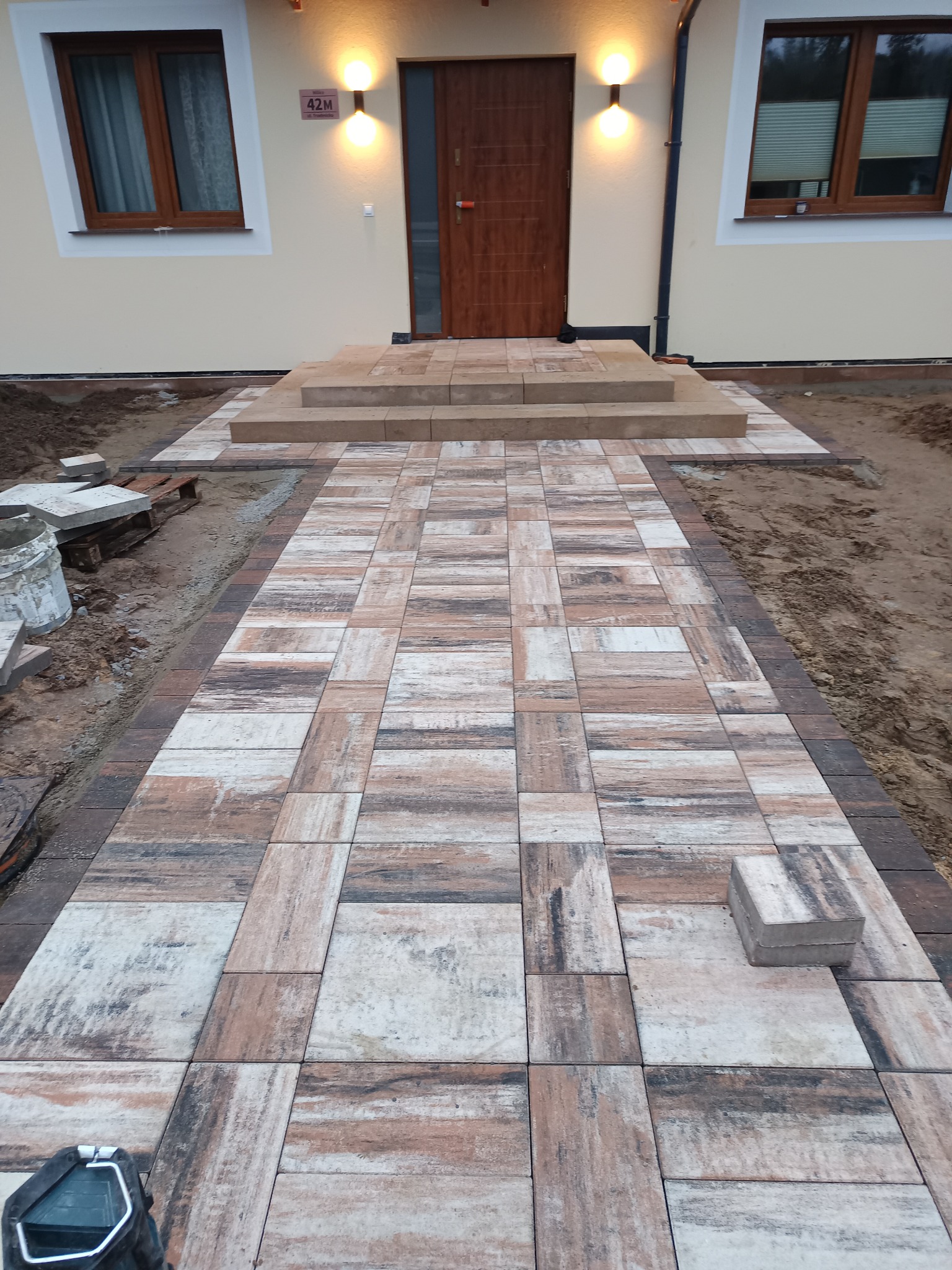 Nowo ułożona ścieżka z kostki brukowej prowadząca do schodów wejściowych domu, z widoczną kostką do ułożenia i narzędziem w rogu kadru.