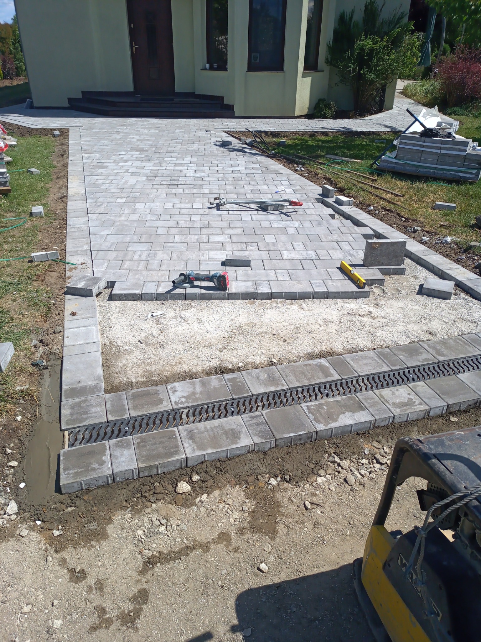 Fragment podjazdu z szarej kostki brukowej w trakcie realizacji, widoczny liniowy odpływ wody, poziomica i wibrator do zagęszczania, dom w tle.