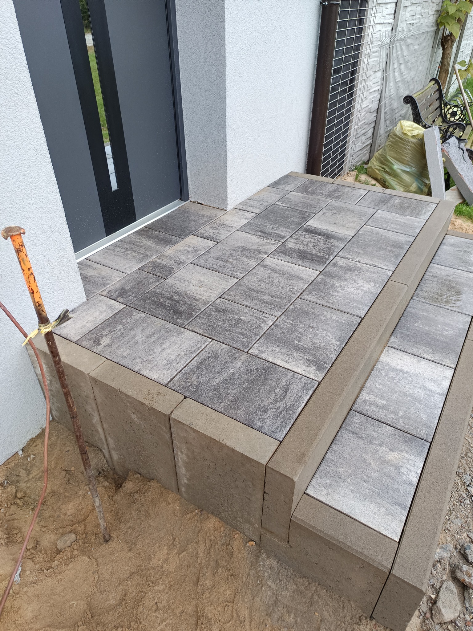 Schody wejściowe wyłożone szarą kostką brukową o nieregularnym wzorze, z widocznym metalowym prętem wbitym w ziemię obok, przy nowoczesnych, ciemnych drzwiach.