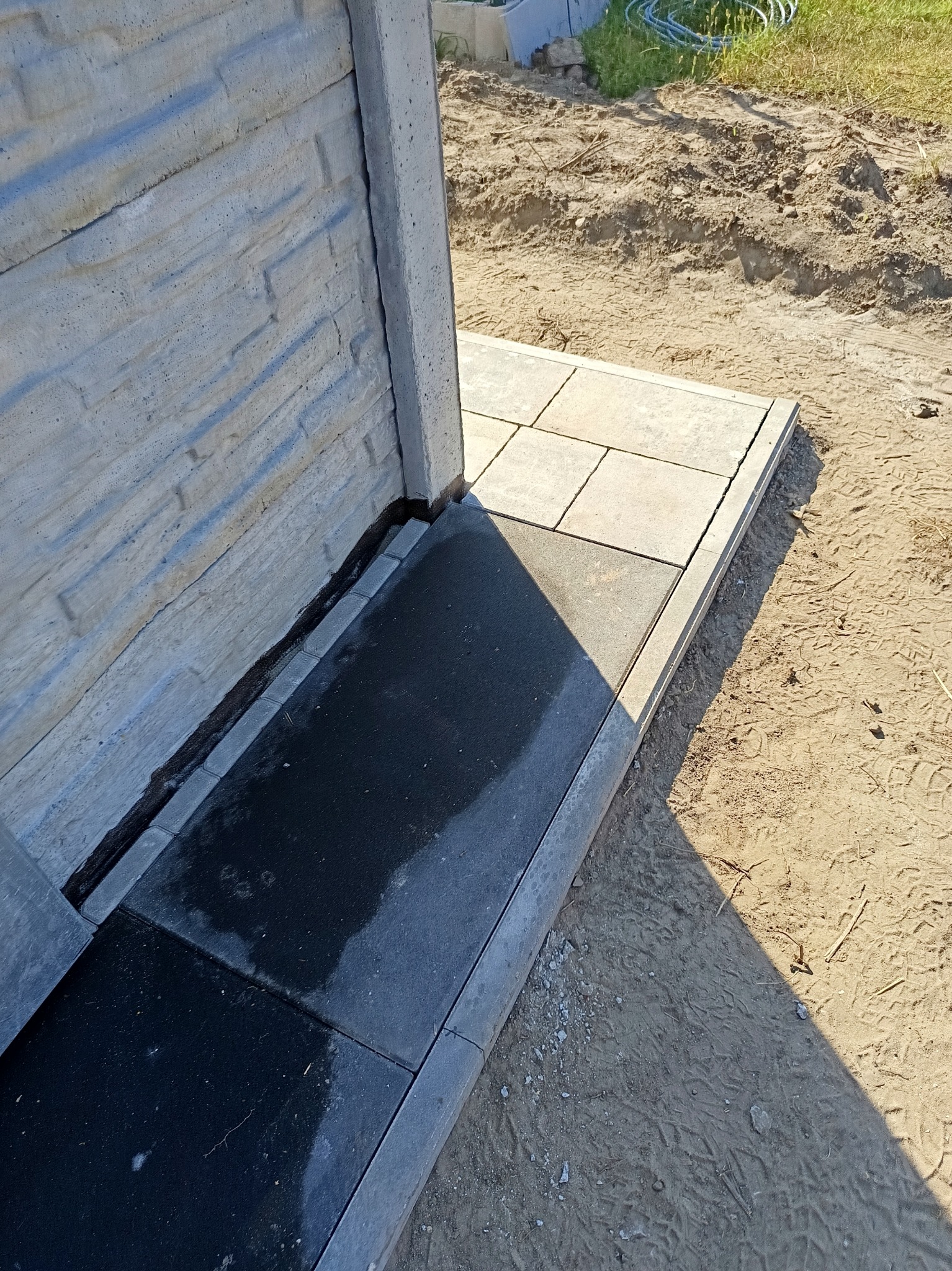 Fragment betonowego ogrodzenia z ułożoną, częściowo mokrą, ciemną kostką brukową i jasnymi płytami chodnikowymi, widoczne ślady po pracy w ziemi.