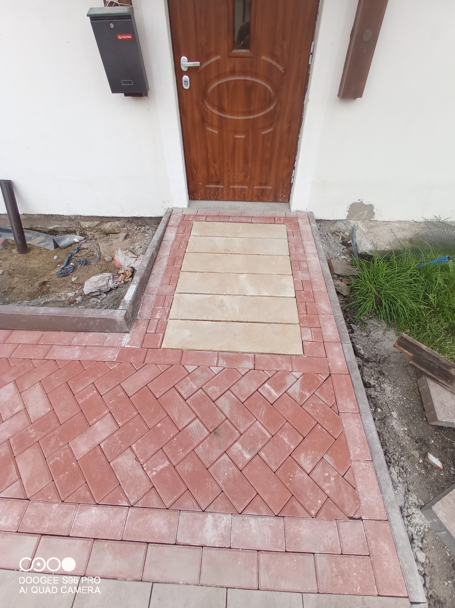 Częściowo ułożony chodnik z czerwonej i beżowej kostki brukowej prowadzący do drewnianych drzwi wejściowych, widoczna skrzynka pocztowa i fragment trawnika