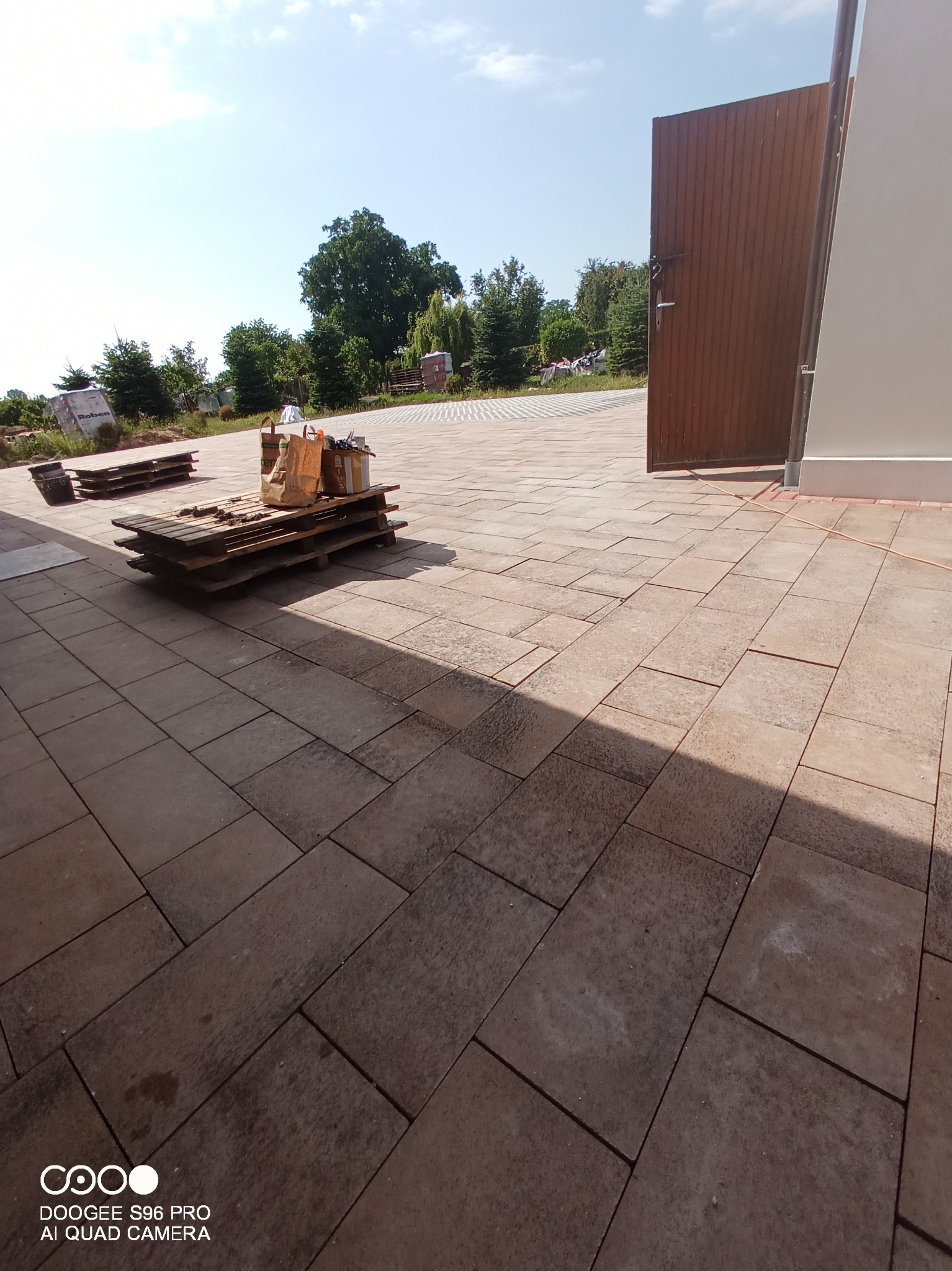 Ułożona kostka brukowa w odcieniach brązu prowadząca do otwartej, drewnianej bramy. Na paletach widoczne materiały budowlane. Częściowo słoneczne oświetlenie.