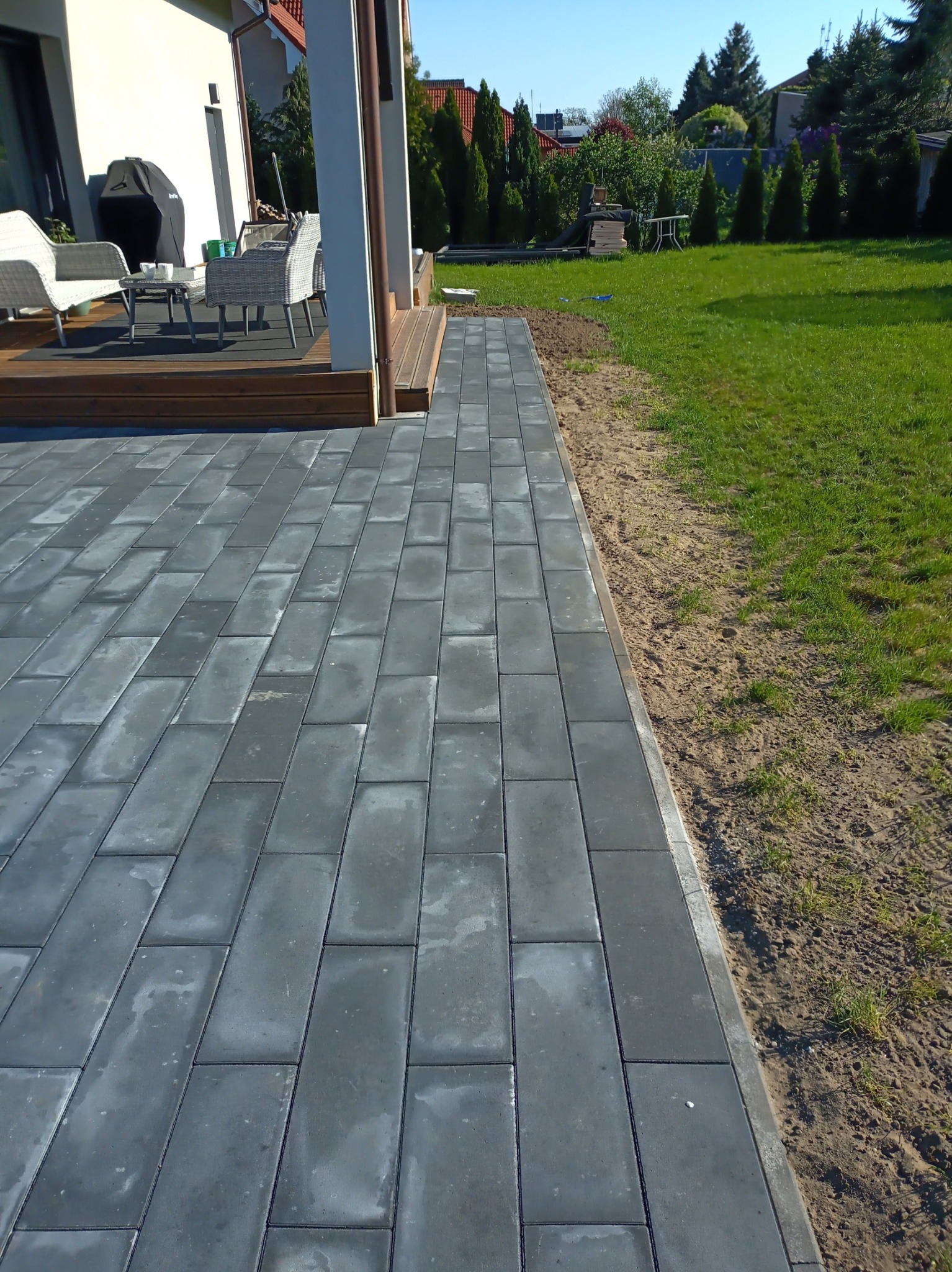 Ułożona szara kostka brukowa na posesji, widoczny fragment tarasu z meblami ogrodowymi, trawnik w tle.