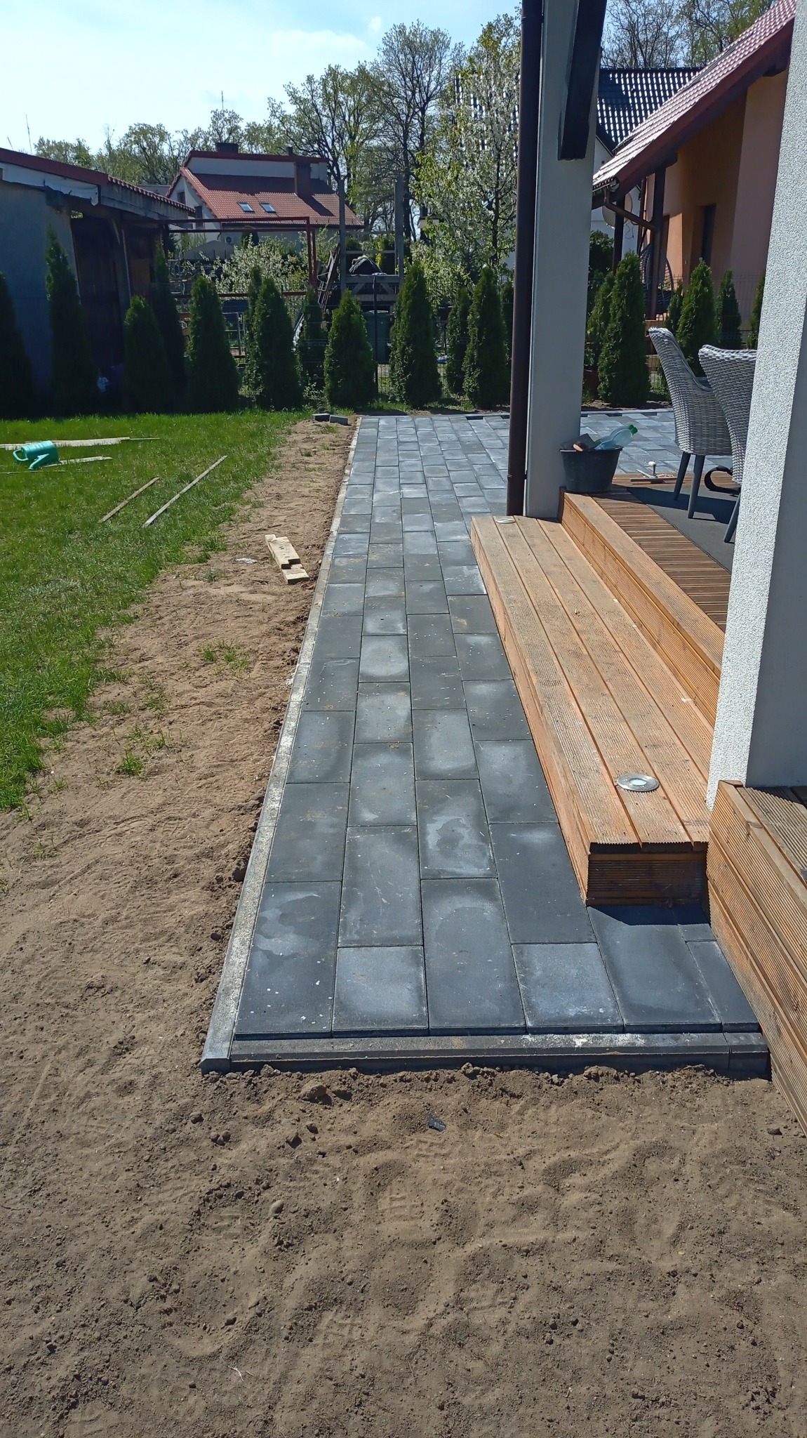 Świeżo ułożona, szara kostka brukowa tworząca ścieżkę wzdłuż drewnianego tarasu, z widocznym piaskowym podłożem i zielonym trawnikiem w tle.