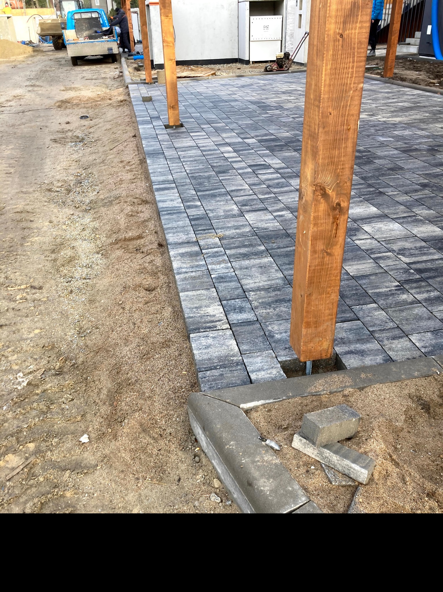Ułożona szara kostka brukowa z drewnianymi słupami w trakcie budowy, widoczne niedokończone krawędzie i piasek, mały niebieski pojazd w tle.