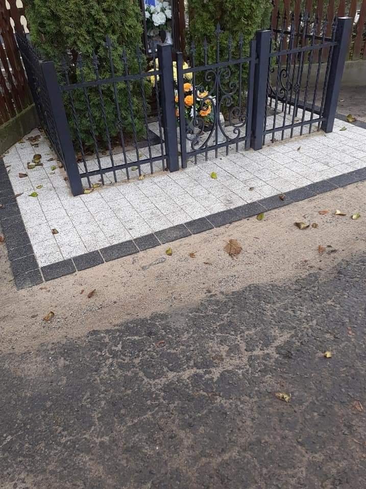 Wykończone miejsce pamięci z jasnoszarą kostką brukową obramowaną ciemniejszą kostką, żelazne ogrodzenie z ozdobnymi elementami, jesienne liście na kostce i ziemi.