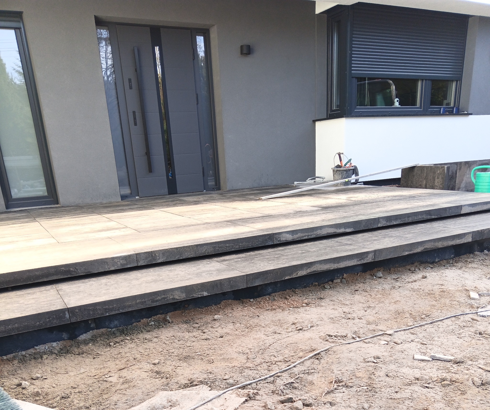 Nowoczesne schody wejściowe z szarej kostki brukowej, częściowo ułożone, z widocznymi narzędziami i elementami wykończeniowymi. Dom w tle.