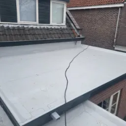 Płaski dach pokryty jasnoszarą membraną, widoczne połączenia arkuszy, z czarnym kablem biegnącym po powierzchni, fragment muru z cegły w tle.