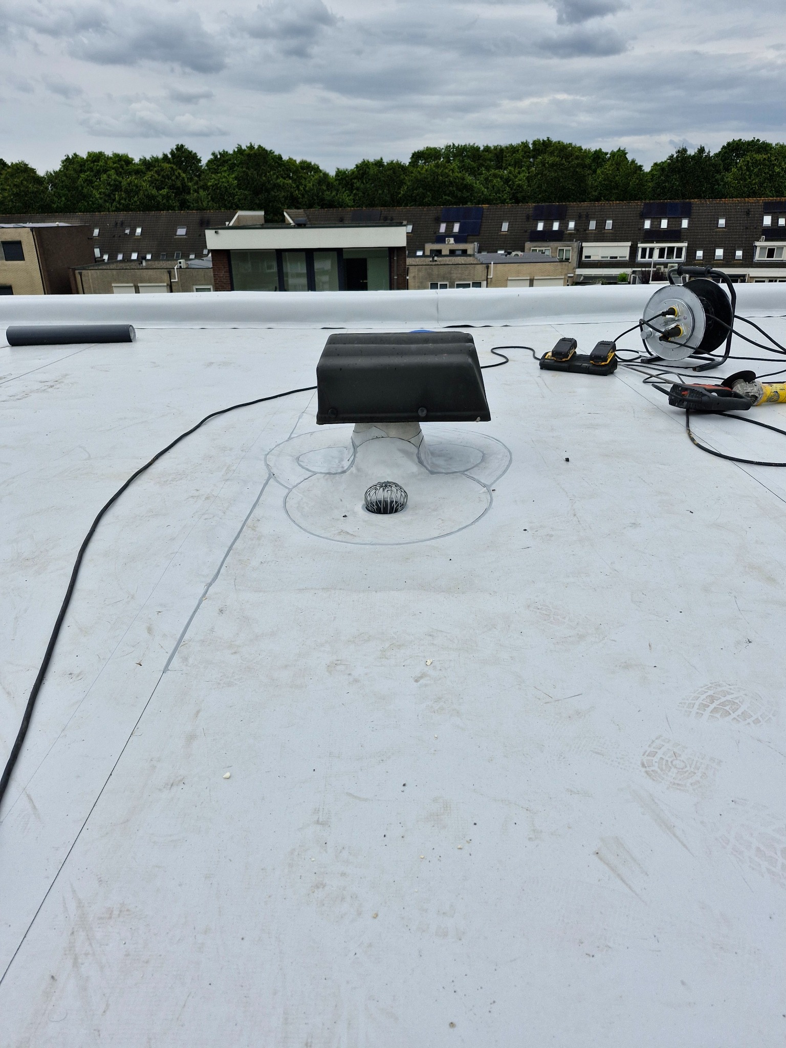 Biała membrana dachowa z zainstalowanym odpowietrznikiem i narzędziami do obróbki blacharskiej, widok z góry na tle zabudowań i zieleni.