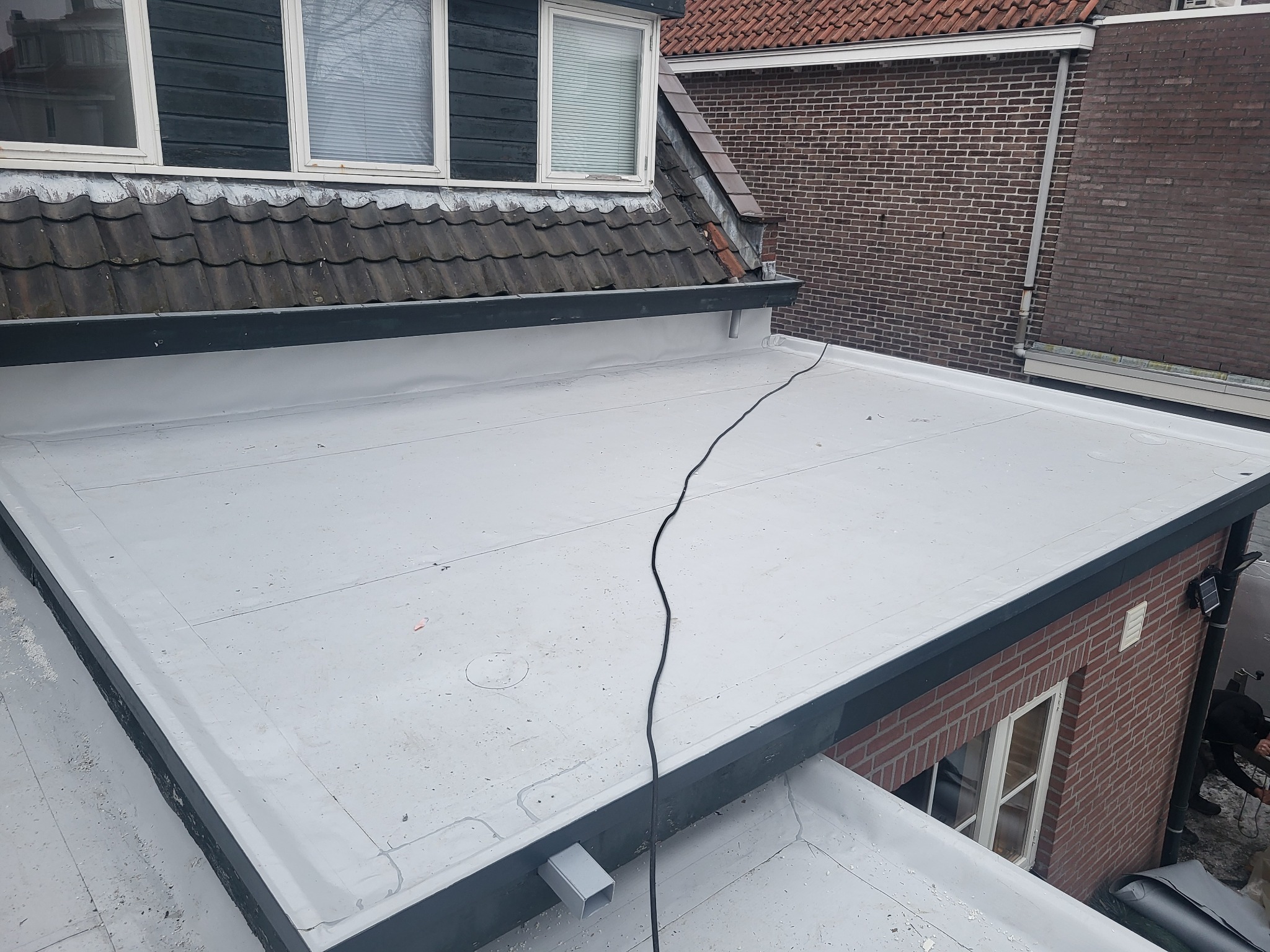 Płaski dach pokryty jasnoszarą membraną, widoczne połączenia arkuszy, z czarnym kablem biegnącym po powierzchni, fragment muru z cegły w tle.