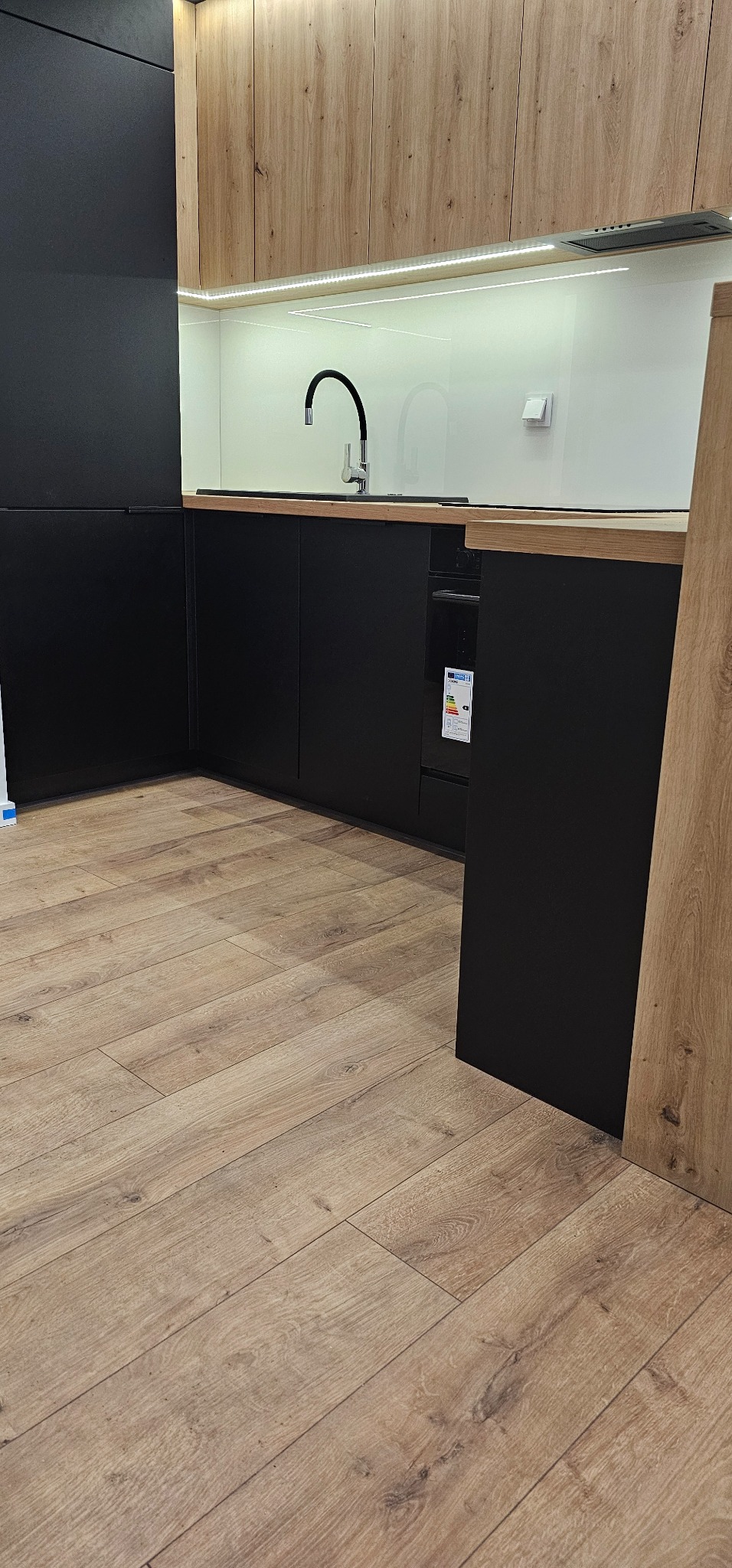 Nowoczesna aranżacja kuchni z czarnymi frontami dolnych szafek i drewnopodobnymi górnymi szafkami, biały panel nad blatem z oświetleniem LED i chromowaną baterią.