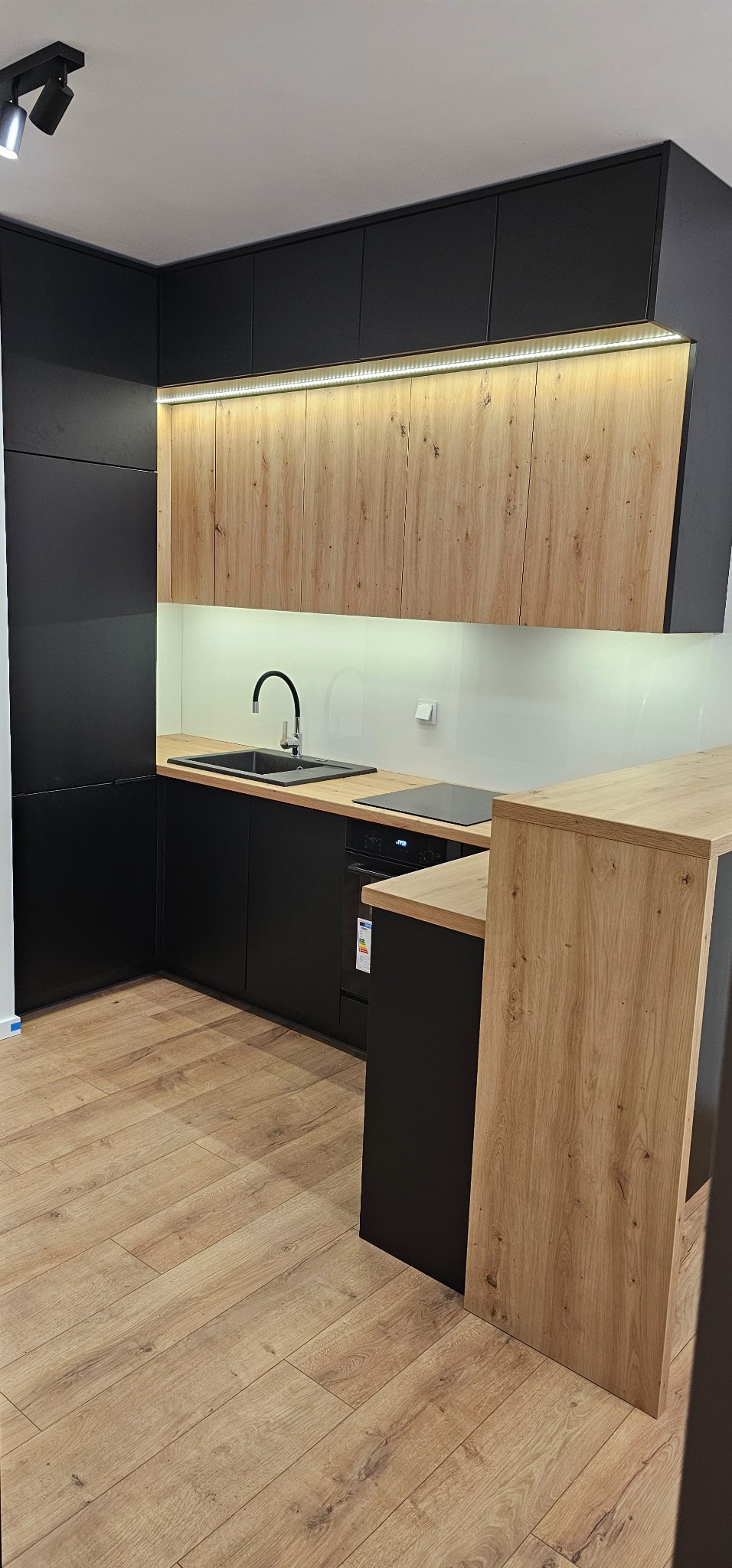 Nowoczesna kuchnia z czarnymi szafkami dolnymi i drewnopodobnymi górnymi, podświetlona listwą LED, z czarnym zlewem i płytą indukcyjną.