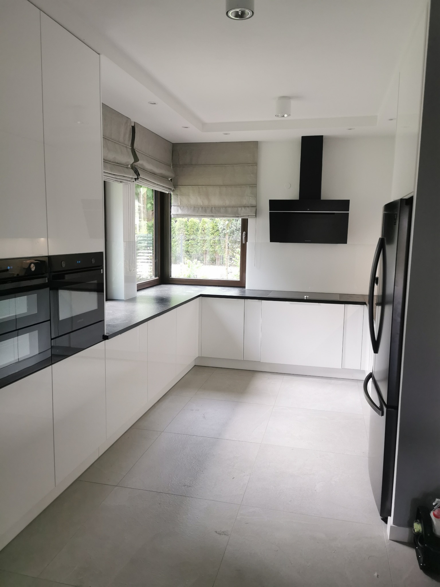 Minimalistyczna, jasna kuchnia z białymi, lakierowanymi szafkami, czarnym blatem i sprzętem AGD, okno z szarą roletą rzymską, szare płytki na podłodze.