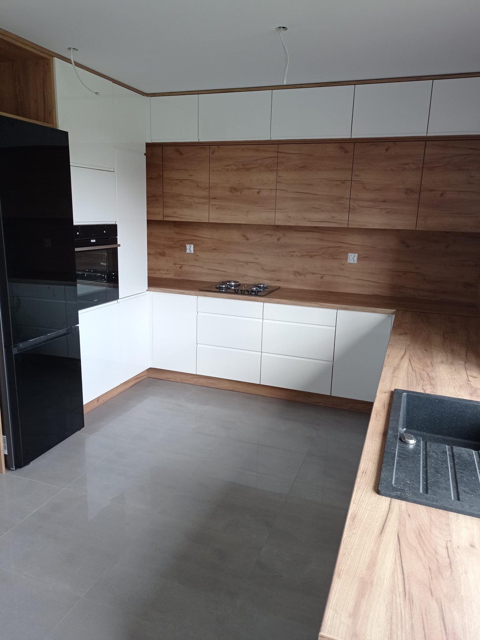 Nowoczesna kuchnia z białymi frontami szafek dolnych i górnych, oraz drewnopodobnymi szafkami górnymi i blatem, czarna lodówka po lewej stronie, szare płytki na podłodze.