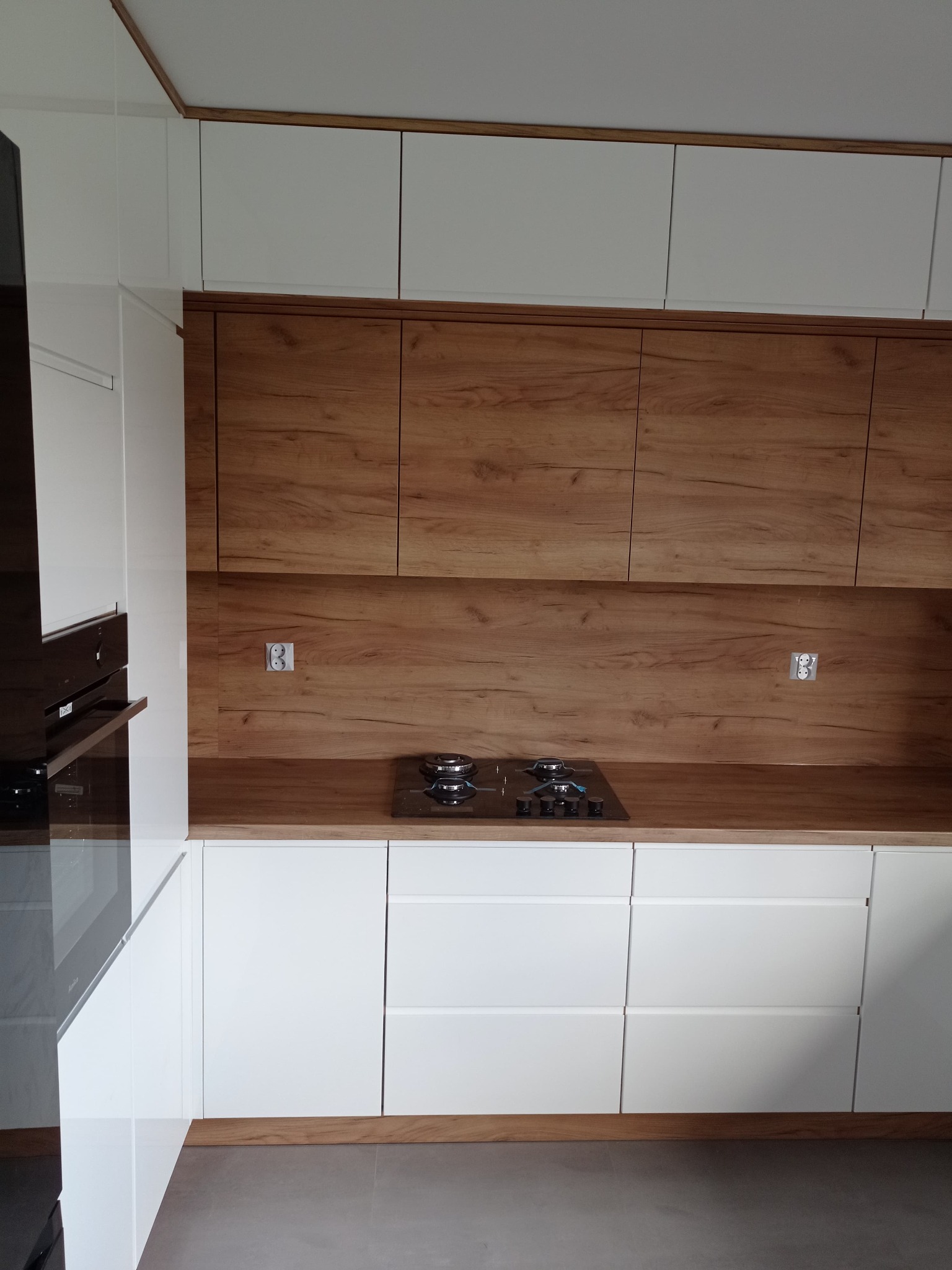 Nowoczesna kuchnia z białymi szafkami dolnymi i górnymi oraz drewnianymi szafkami wiszącymi i panelem nad blatem, płyta gazowa na blacie, widoczne gniazdka elektryczne na panelu.