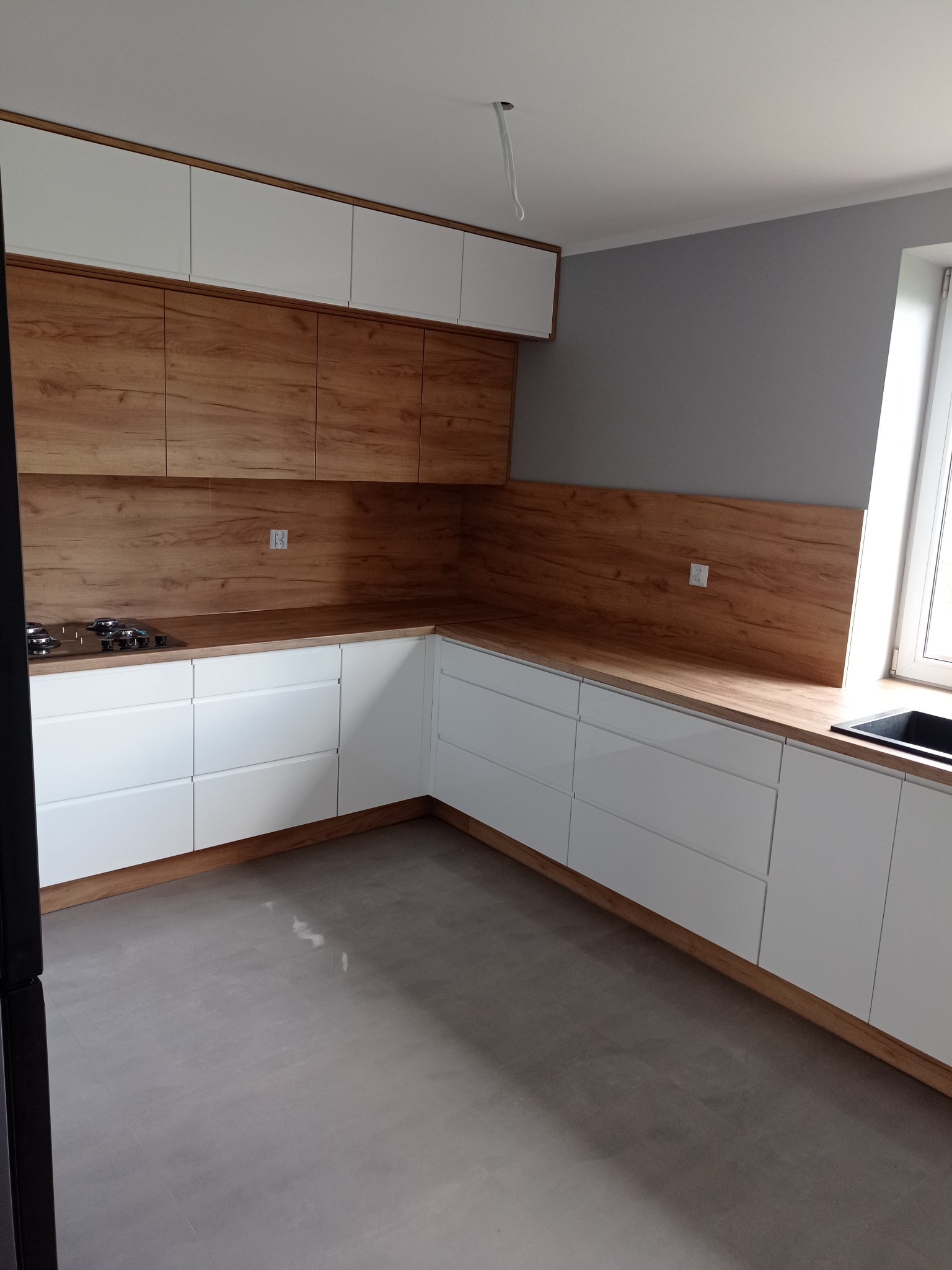 Jasna kuchnia z białymi szafkami i drewnianymi blatami oraz panelami ściennymi, widoczna płyta grzewcza i zlew, szare ściany, minimalistyczny styl.