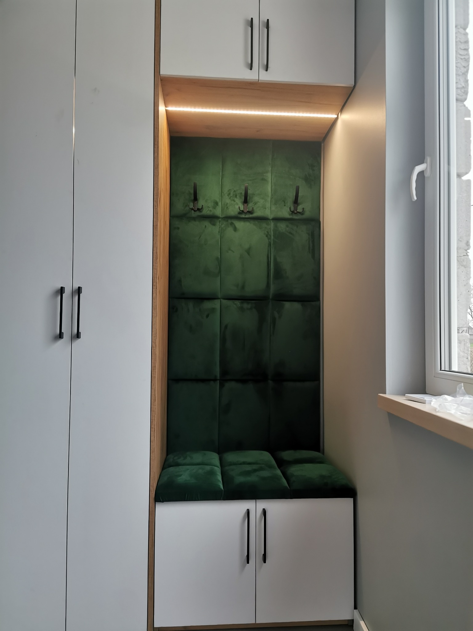 Wnęka z siedziskiem i wieszakami, obok biała szafa z czarnymi uchwytami, oświetlenie LED, zielona tapicerka w kwadraty, widok na okno.