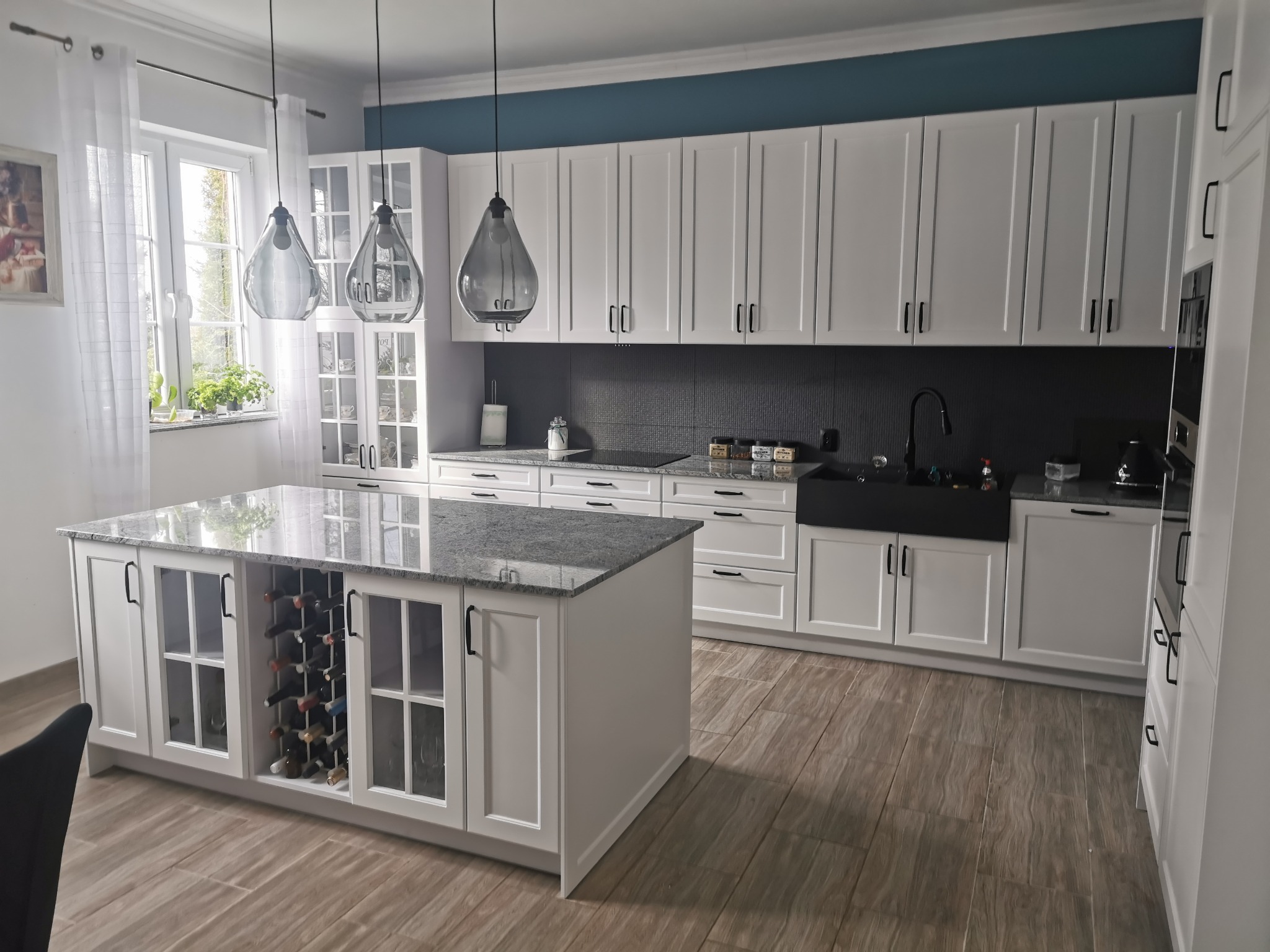 Jasna kuchnia z białymi szafkami, czarnymi uchwytami i wyspą z marmurowym blatem oraz wbudowanym regałem na wino, oświetlona wiszącymi lampami w kształcie kropli.