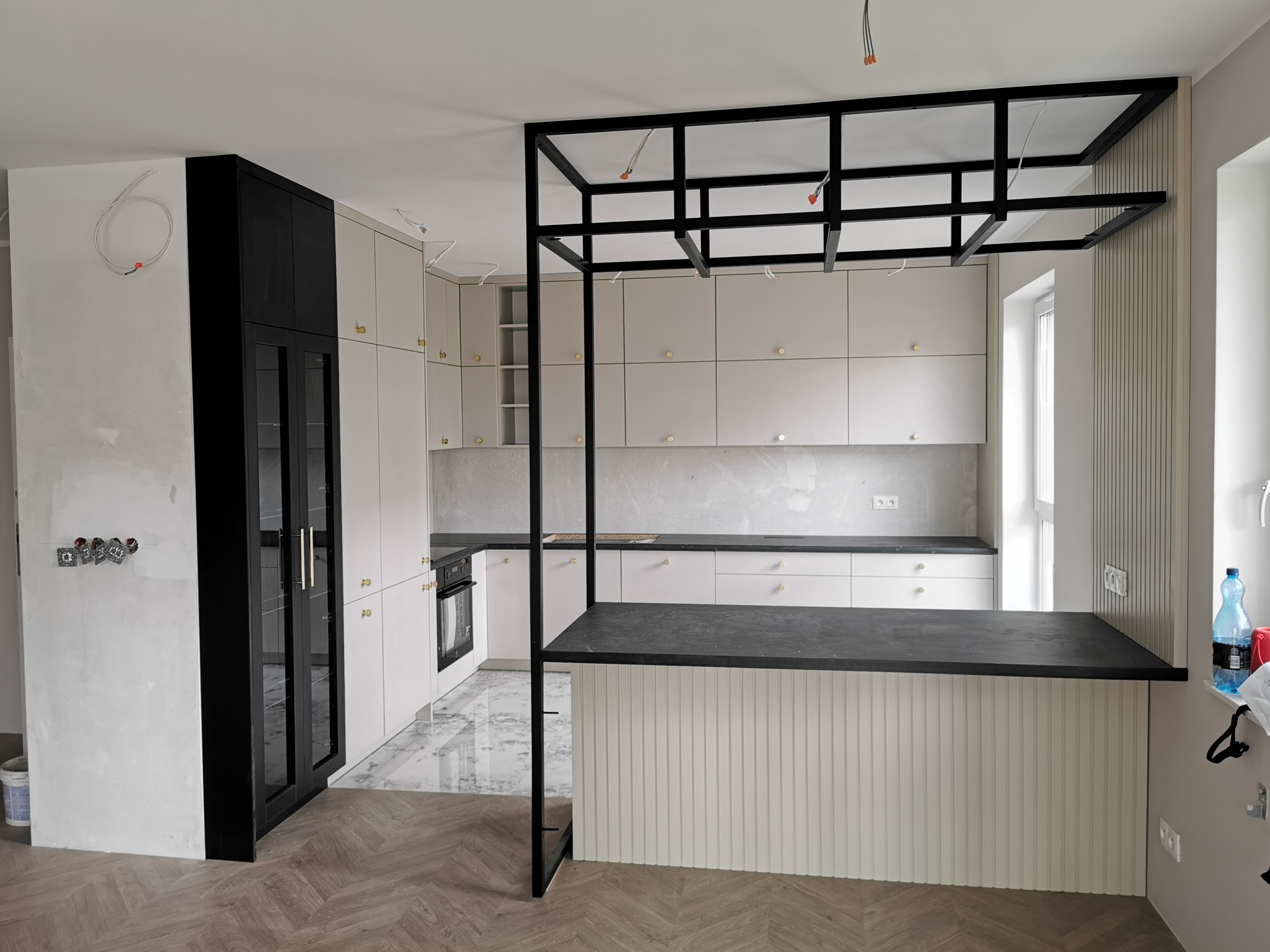 Wnętrze kuchni w trakcie wykańczania z kremowymi szafkami, czarnym regałem nad wyspą i wykończonymi podłogami w jodełkę.