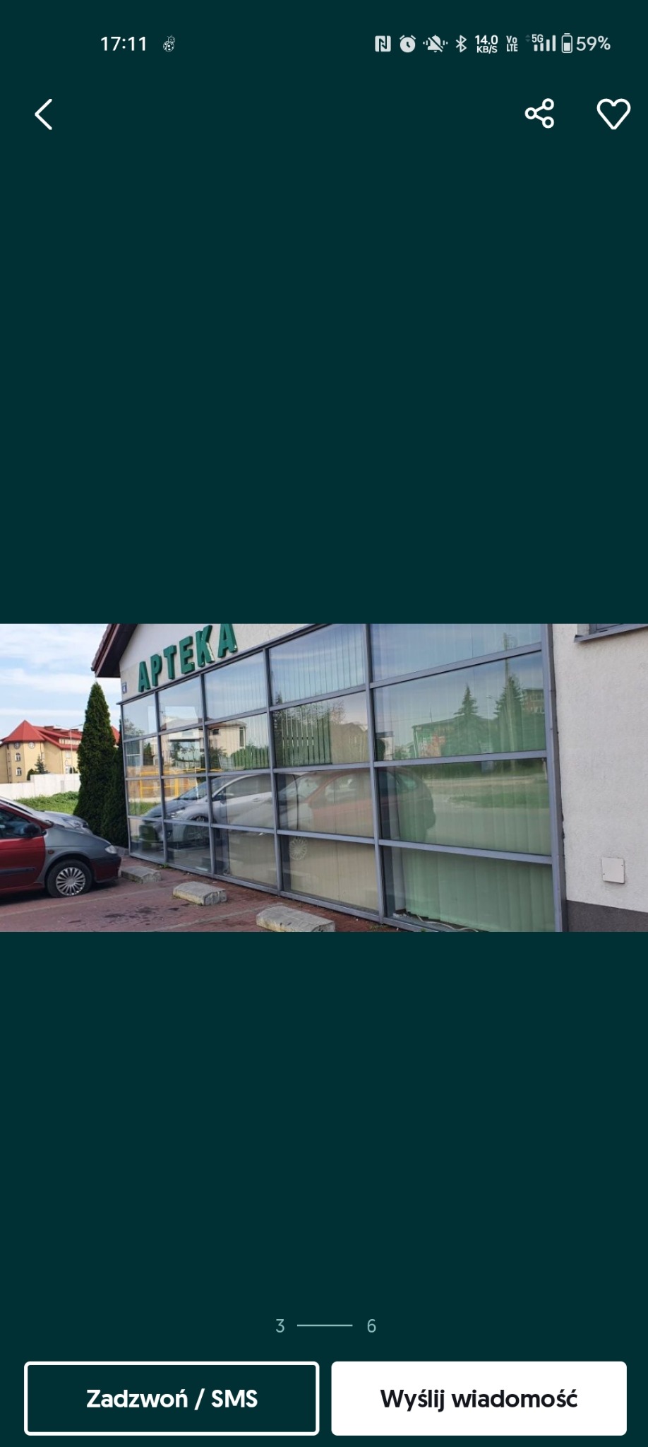 Zewnętrzny widok witryny apteki z dużymi oknami, w których odbijają się budynki i drzewa, z zaparkowanymi samochodami przed nią.