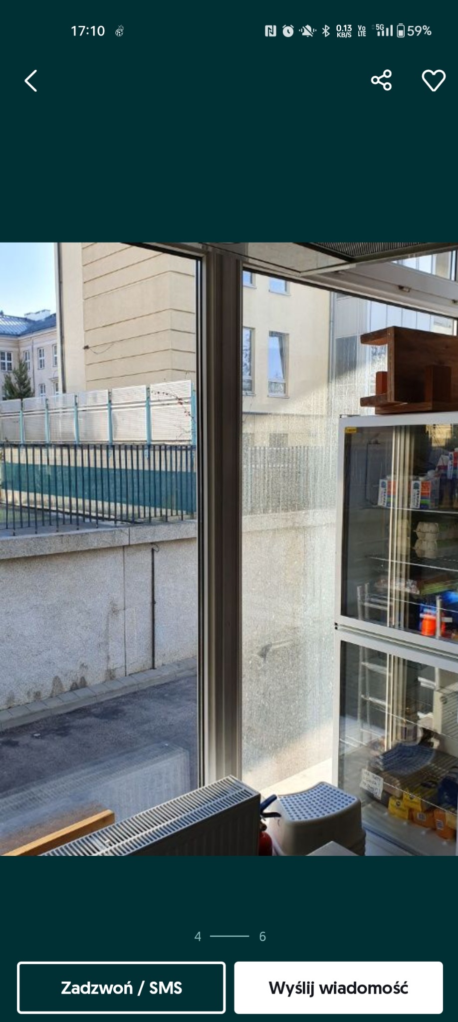 Widok z okna na budynek i balkon z barierką. Szyba okna jest brudna, widoczne zacieki. Wewnątrz pomieszczenia widoczny grzejnik, stołek i regał z produktami.