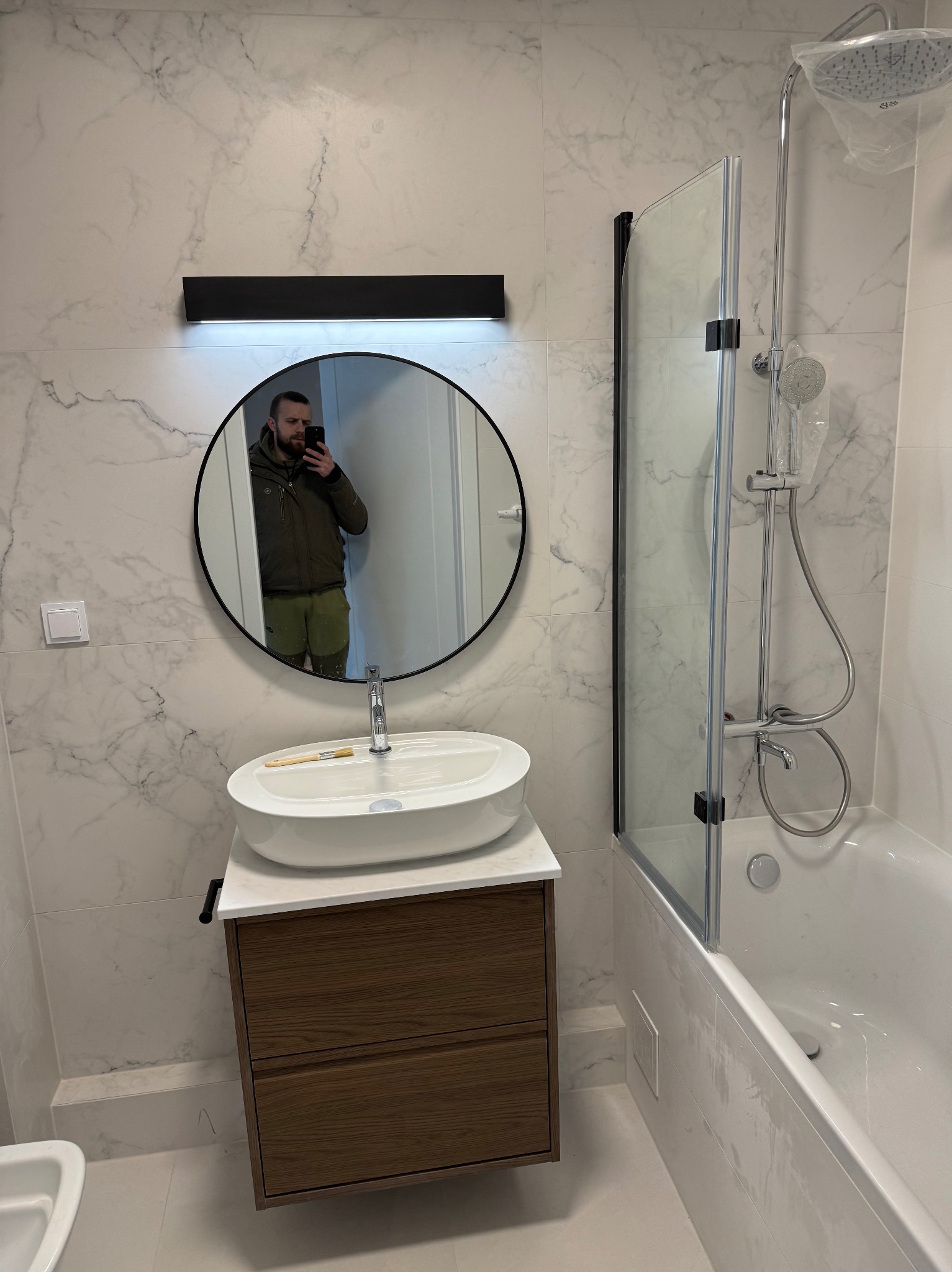 Nowoczesna łazienka z marmurowymi płytkami, owalną umywalką nablatową, drewnianą szafką pod umywalką, czarnym oświetleniem nad lustrem i kabiną prysznicową z osłoną, w lustrze odbija się osoba...