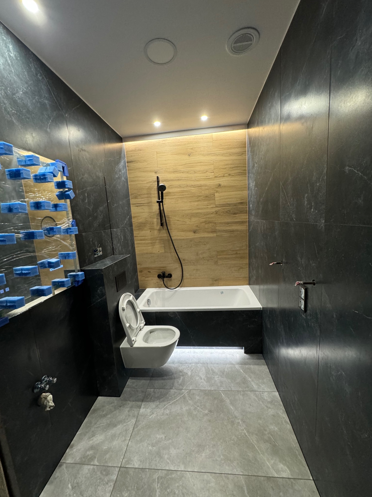 Nowoczesna łazienka z czarnymi marmurkowymi płytkami na ścianach, drewnopodobnymi płytkami w strefie wanny, podwieszaną toaletą z otwartą deską, szarą podłogą i oświetleniem LED. Fragment ściany...