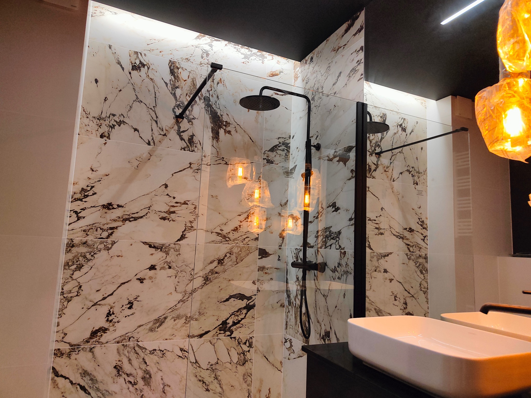 Wykończona kabina prysznicowa typu walk-in z czarną armaturą i marmurowymi płytkami na ścianach, oświetlona dekoracyjną lampą wiszącą z żarówkami Edisona, widoczna biała umywalka nablatowa.