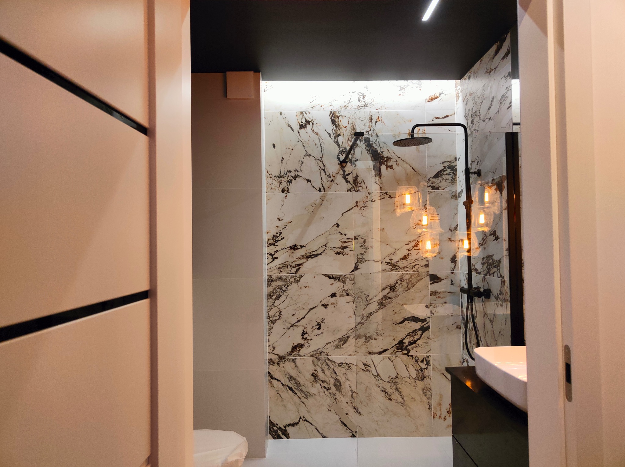 Nowoczesna łazienka z marmurowymi płytkami, czarną armaturą i podwieszanymi lampami żarówkami, widok zza uchylonych drzwi.
