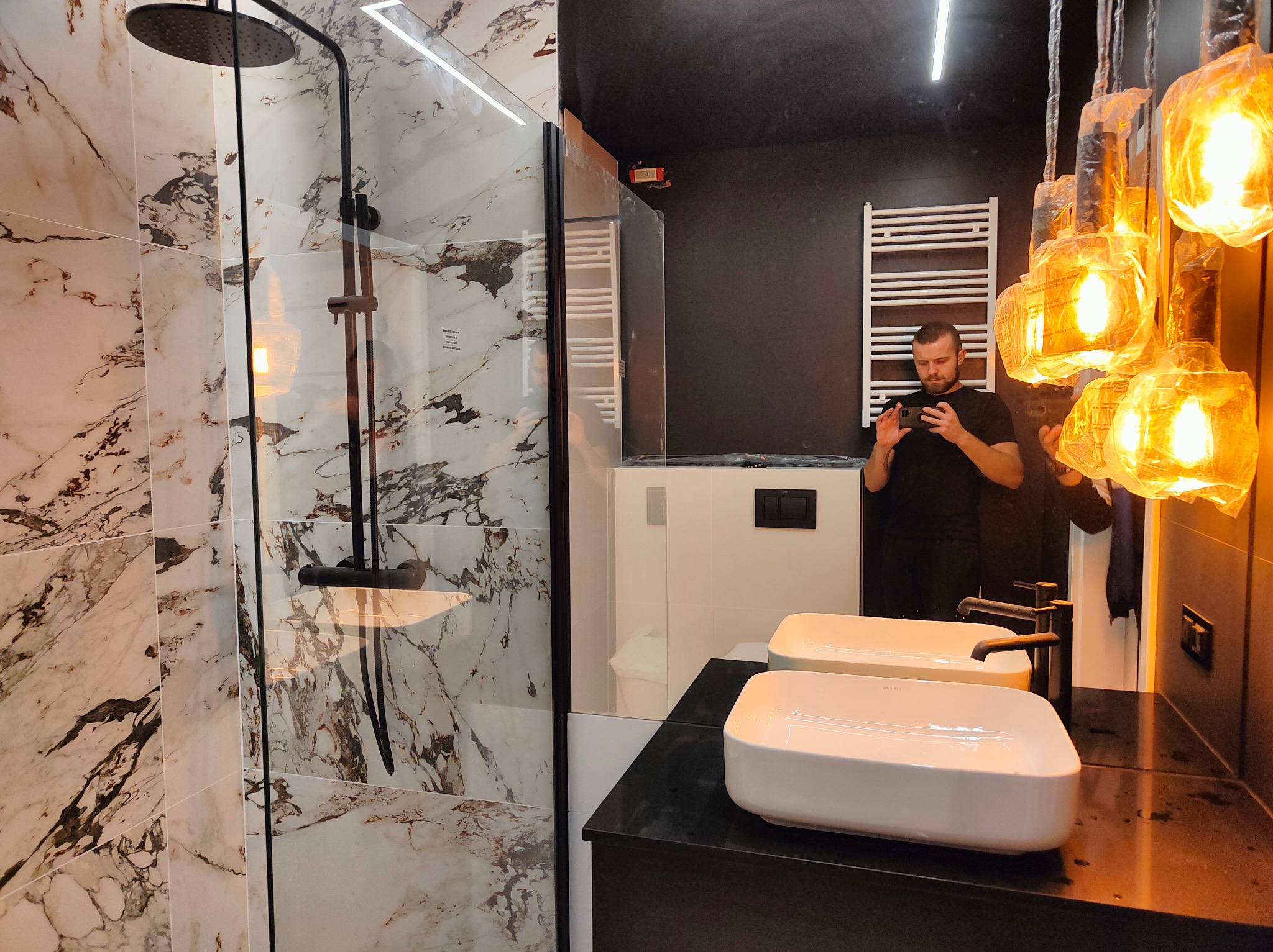 Nowoczesna łazienka z kabiną prysznicową typu walk-in, wyłożona płytkami imitującymi marmur. Widoczne dwie białe umywalki nablatowe, czarna armatura i wiszące lampy w stylu industrialnym. Mężczyzna...