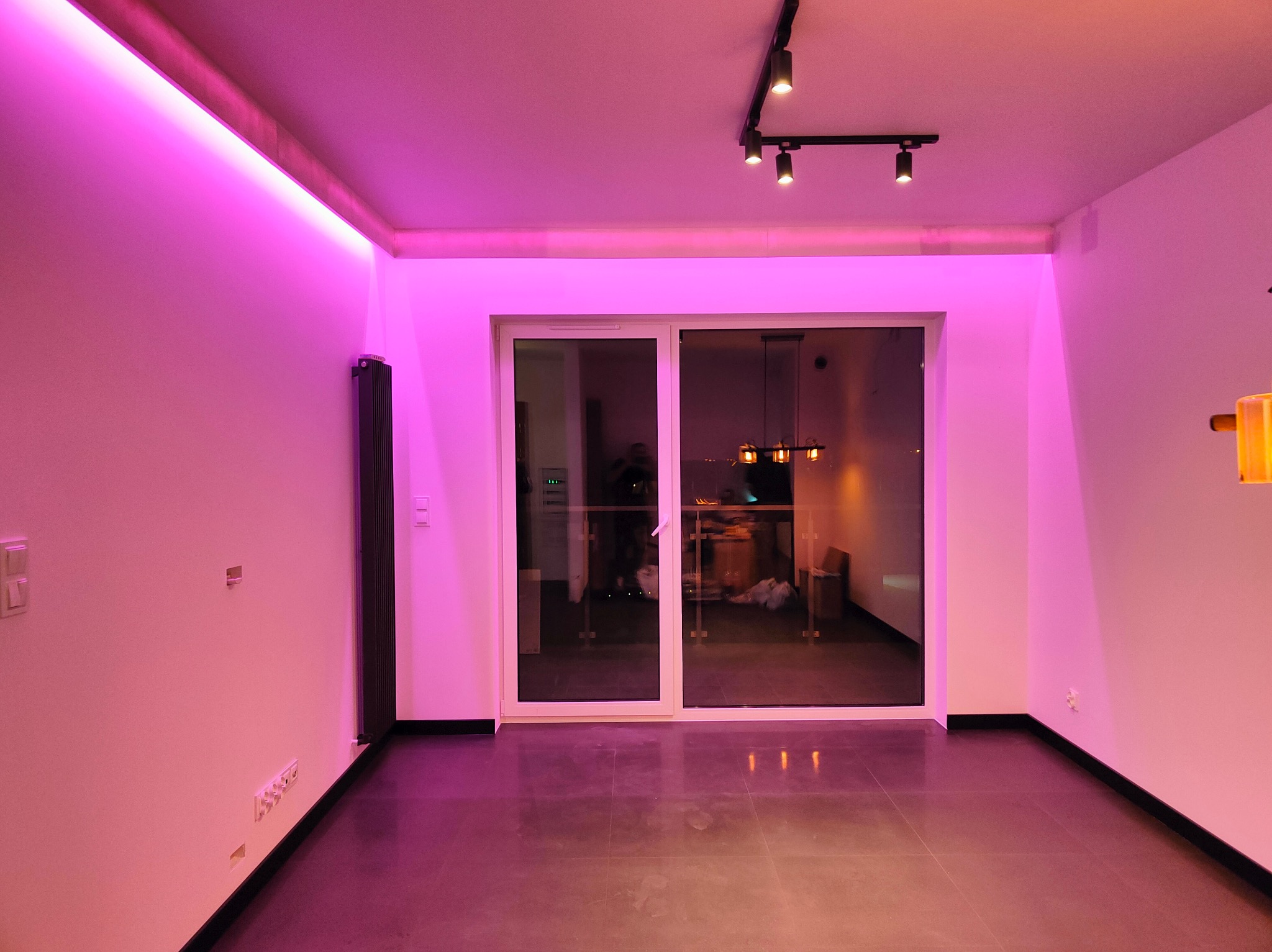 Pusty pokój z szarymi płytkami podłogowymi i białymi ścianami oświetlonymi intensywnym różowym światłem LED na suficie, widoczne przesuwne drzwi balkonowe i czarne reflektory sufitowe.