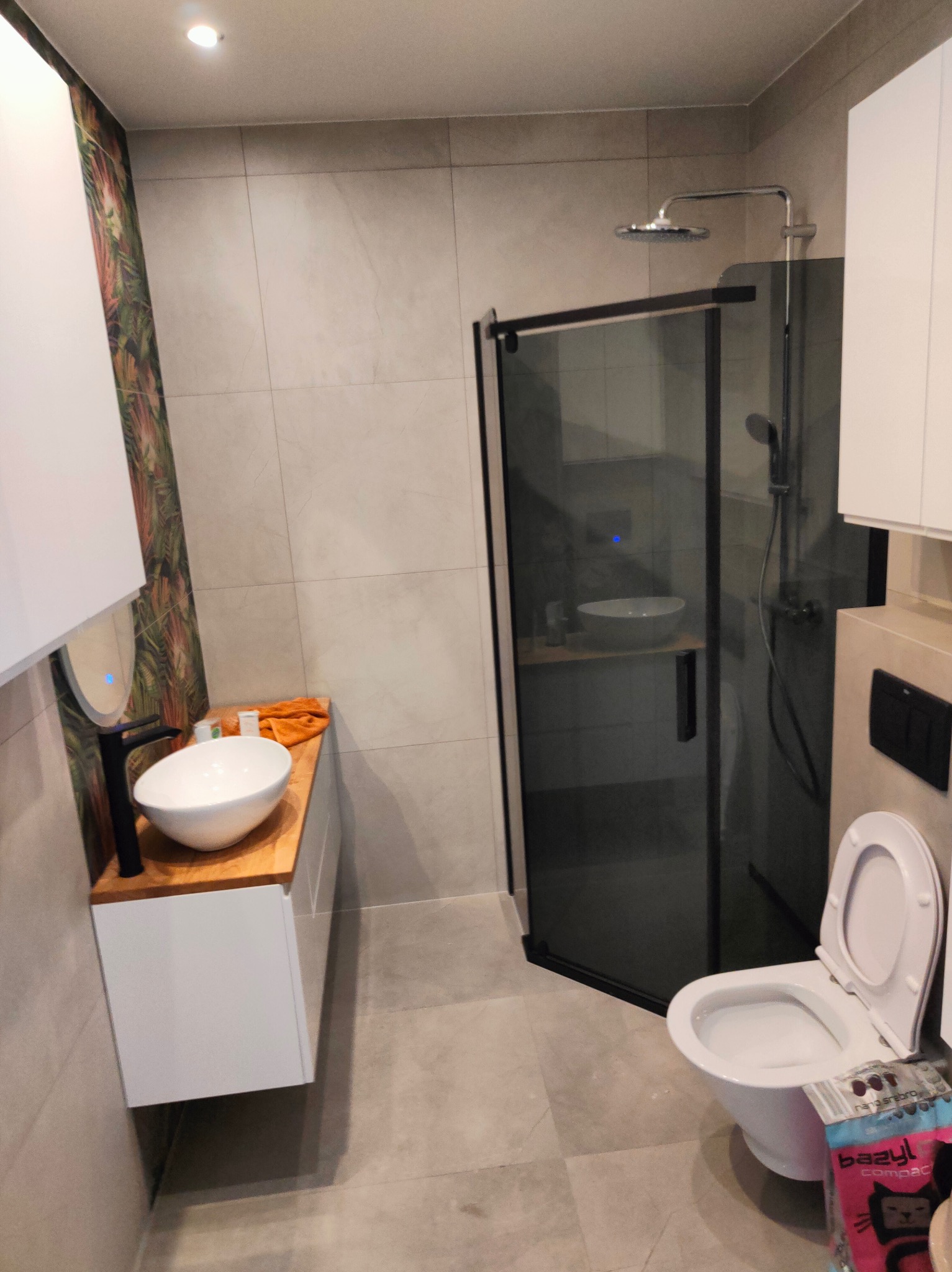 Nowoczesna łazienka z kabiną prysznicową typu walk-in z czarnymi ramami, umywalką nablatową na drewnianym blacie, wiszącą toaletą i akcentem w postaci tapety w tropikalne liście.