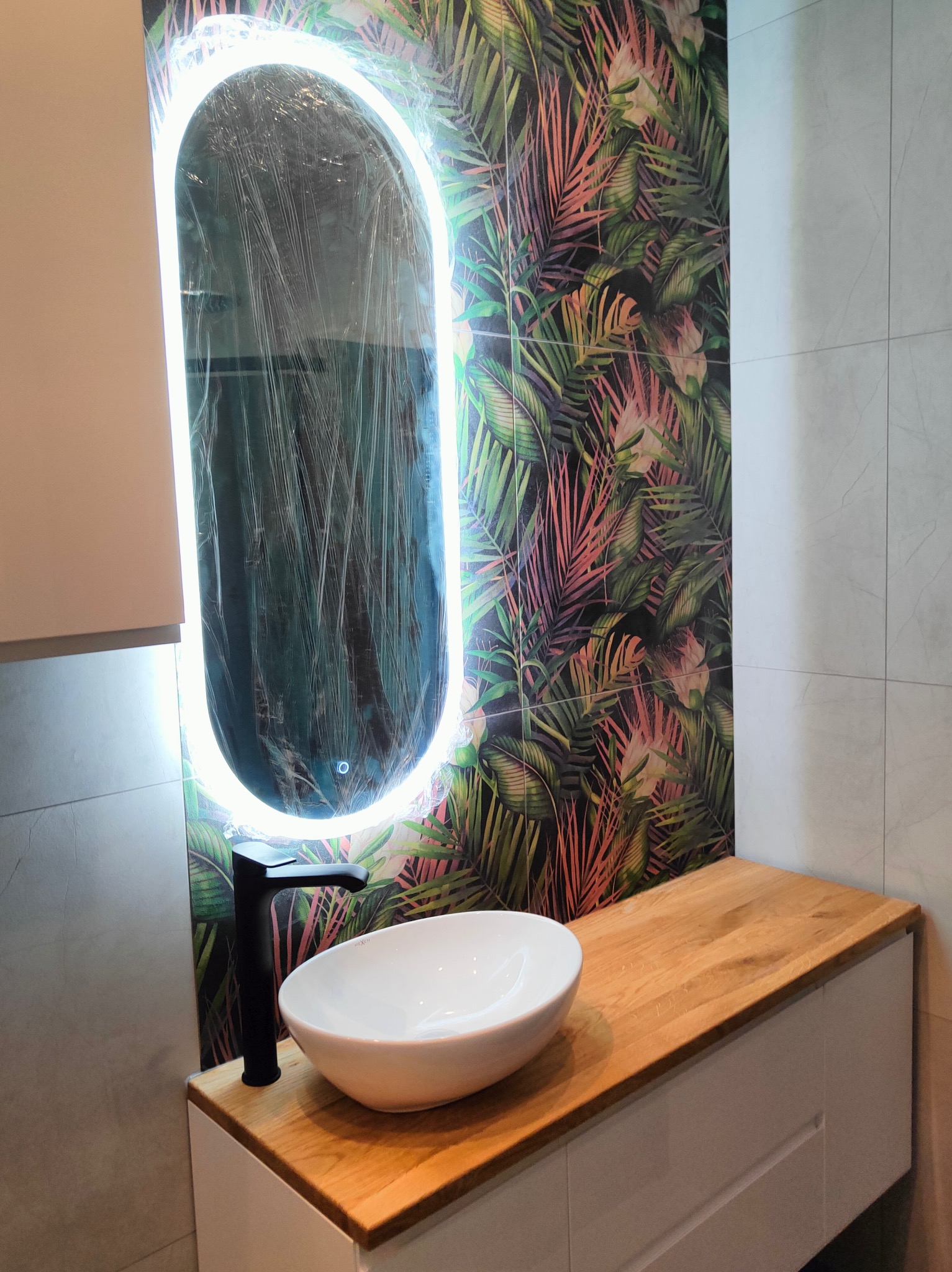 Nowoczesna łazienka z umywalką nablatową, czarną armaturą i podświetlanym lustrem owiniętym folią ochronną na tle ściany w tropikalne liście.