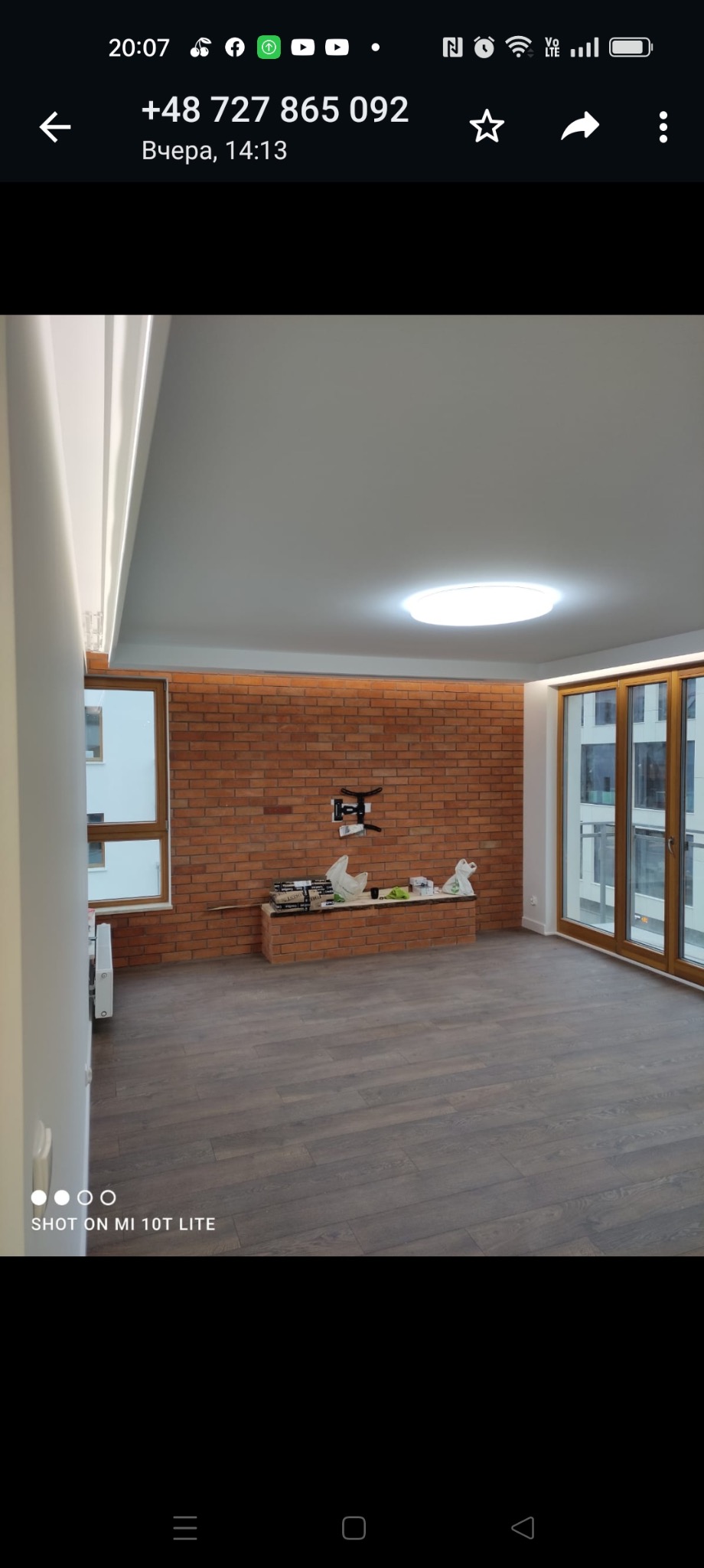 Wnętrze mieszkania w trakcie wykańczania: ściana z czerwonej cegły, zamontowany uchwyt na telewizor, podłoga wyłożona panelami imitującymi drewno, duże okna balkonowe z drewnianymi ramami, widoczne...