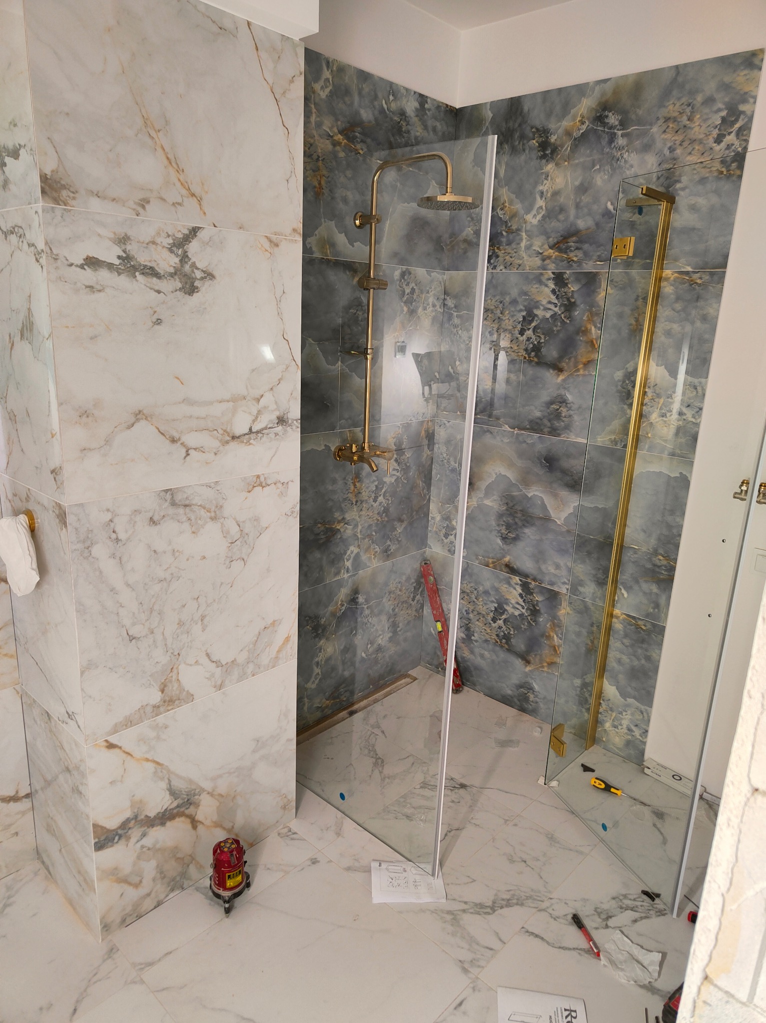 Montaż kabiny prysznicowej ze złotymi elementami w łazience w Gdańsku, widoczne narzędzia i laserowy poziomica, marmurowe płytki na ścianach i podłodze.