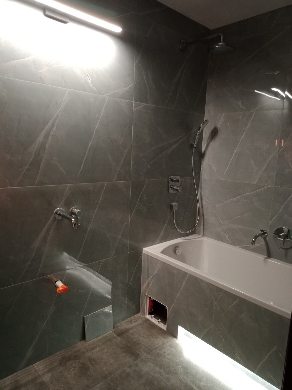Wnętrze łazienki z szarymi płytkami imitującymi marmur, białą wanną z podświetleniem LED u dołu, zamontowaną armaturą i niezakończonymi pracami wykończeniowymi przy podłączeniu rur oraz brakiem...