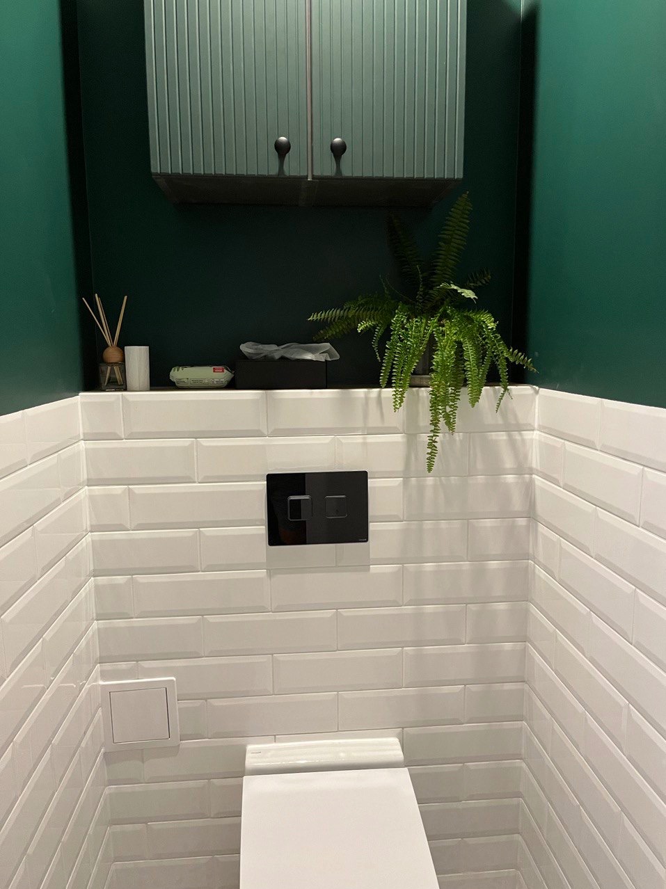 Nowoczesna toaleta z białymi, prostokątnymi płytkami, ciemnozieloną ścianą, szafką z ryflowanymi drzwiczkami i czarnym przyciskiem spłuczki, uzupełniona zieloną paprotką.