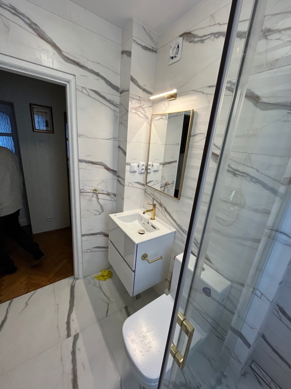 Nowoczesna łazienka z białymi płytkami imitującymi marmur, wisząca szafka z umywalką i złotą baterią, lustro w złotej ramie, kabina prysznicowa z złotymi uchwytami, widoczny fragment toalety...