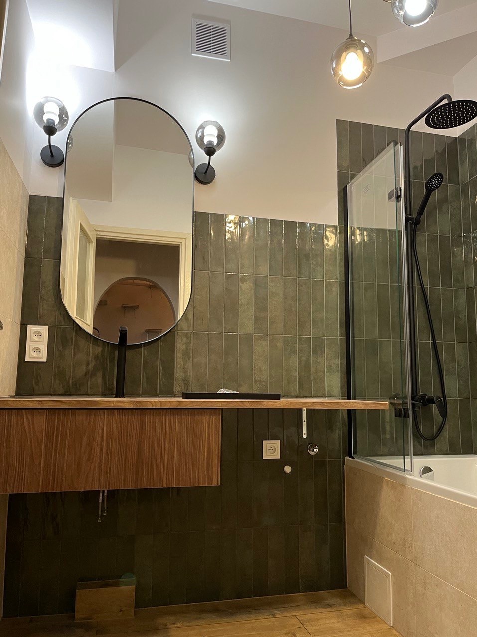 Nowoczesna łazienka z zielonymi, pionowo ułożonymi płytkami, drewnianą szafką pod umywalką z czarną armaturą, owalnym lustrem i kabiną prysznicową z czarnymi elementami.