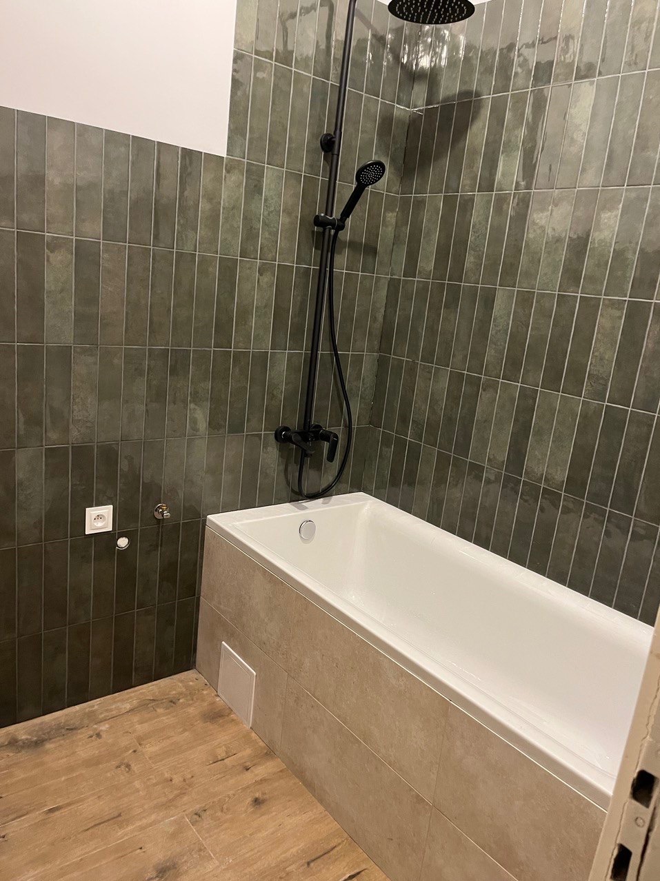 Nowoczesna łazienka z wanną obudowaną płytkami w odcieniach beżu, ściany wyłożone pionowymi, zielonymi płytkami, czarna armatura prysznicowa, drewnopodobna podłoga.