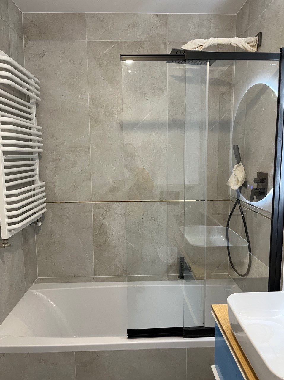 Nowoczesna łazienka z wanną, przeszklonym panelem prysznicowym z czarnymi ramami, grzejnikiem drabinkowym i okrągłym lustrem z podświetleniem LED.