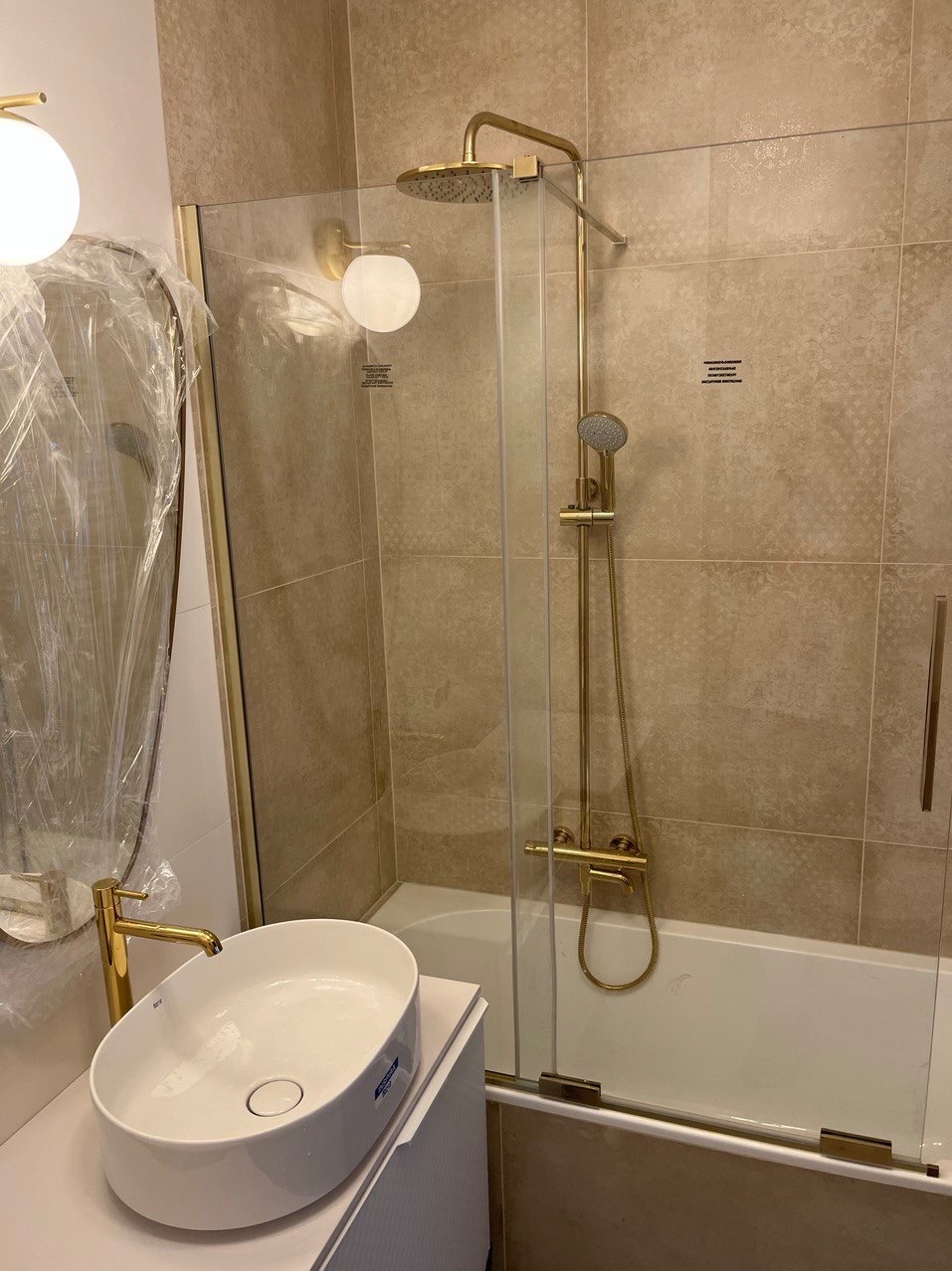 Nowoczesna łazienka z beżowymi płytkami, złotą armaturą, owalną umywalką nablatową i szklanym parawanem nawannowym; lustro owinięte folią ochronną.
