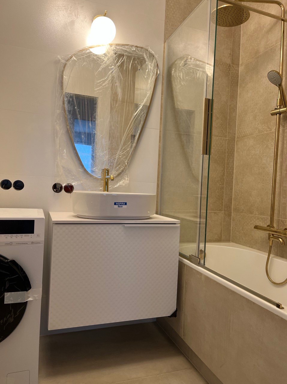 Nowoczesna łazienka w trakcie wykończenia: umywalka nablatowa marki Inspira Roca na szafce z subtelnym wzorem, złota armatura, lustro w folii ochronnej i kabina prysznicowa ze złotymi elementami.